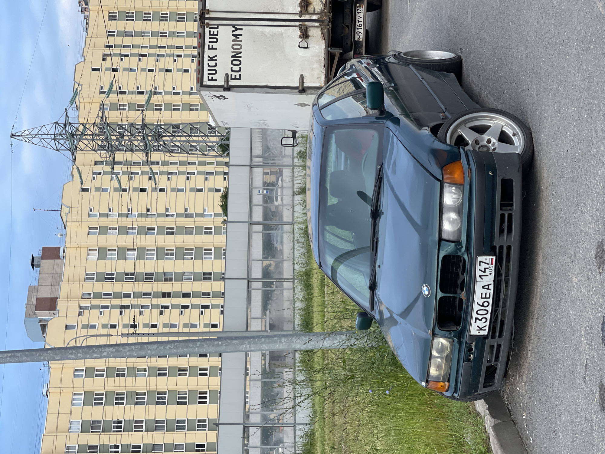 In an urban environment, a dark green BMW sedan is parked on the right side of a paved road. The car's front bumper is damaged, and the grille is missing. The license plate reads "K306EA 147 RUS". In the background, a large, light yellow apartment building with green accents dominates the scene. To the right of the BMW, a white box truck is parked, with the words "FUCK FUEL ECONOMY" painted on its side. A power pylon stands tall between the car and the building, with thick cables extending across the sky. The ground on the left side of the car is covered in tall green grass with a few small purple flowers. The sky is overcast with clouds.