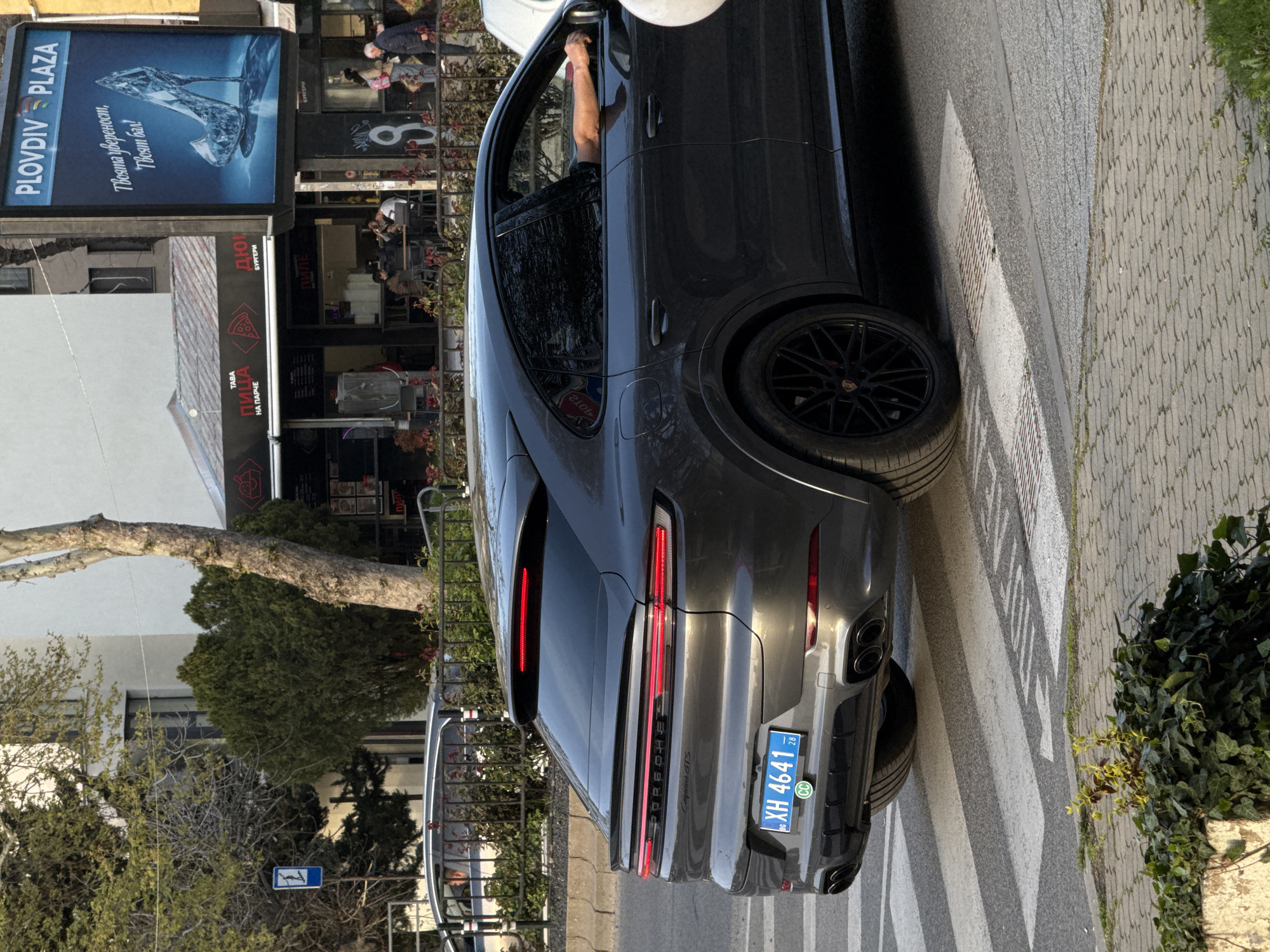 A gray Porsche Cayenne GTS is parked on a crosswalk. The license plate is "XH 4641 28" and has "BG" and "CC" printed on it. The background shows the Plovdiv Plaza sign above a restaurant. People are sitting at the restaurant tables. The street is lined with trees.