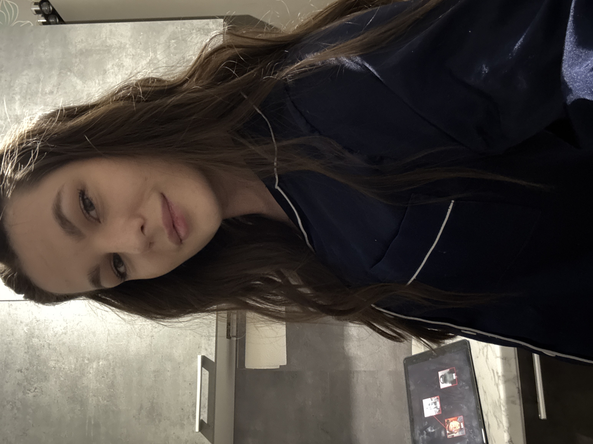 A woman with long brown hair is wearing a navy blue pajama top with white piping. She is positioned on the left side of the frame. To the right, a tablet is visible with multiple images on its screen. The background appears to be a bathroom with a grey wall and a sink. The location may be Chaikovsky, Russia, but there are no visual cues to confirm this.