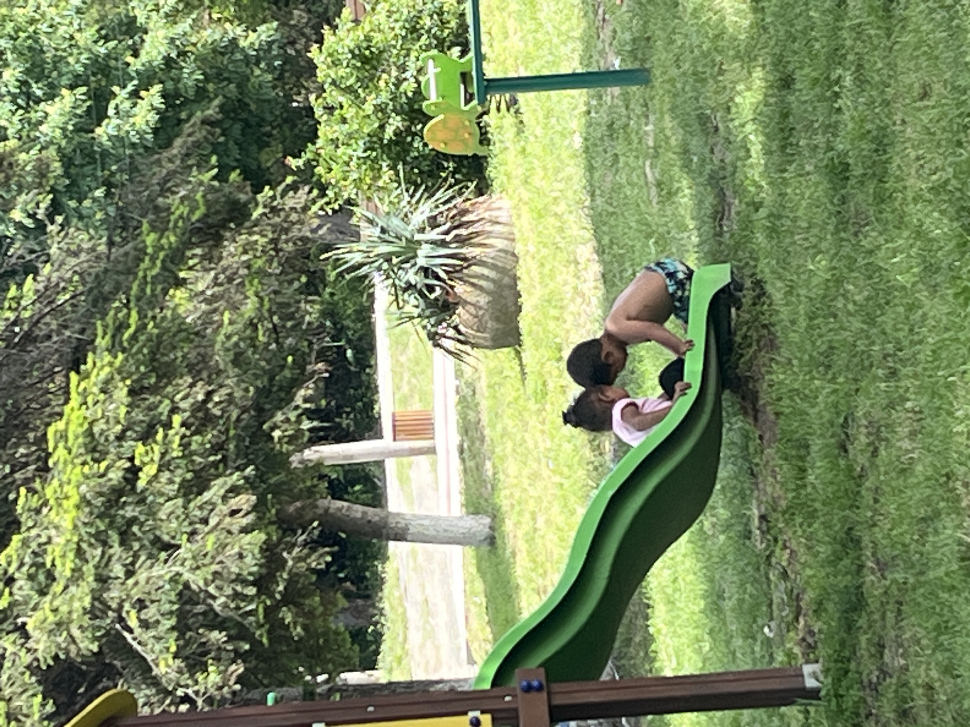 The image shows a young boy and girl on a green slide in Lozarevo, Bulgaria. The boy is shirtless and wearing patterned shorts, positioning himself at the top of the slide. The girl, wearing a light pink shirt, is lying on the slide, facing upwards. The slide is situated in a grassy area with trees and bushes in the background. A green metal structure with a turtle-shaped figure is visible near the slide. Another playground structure is visible.