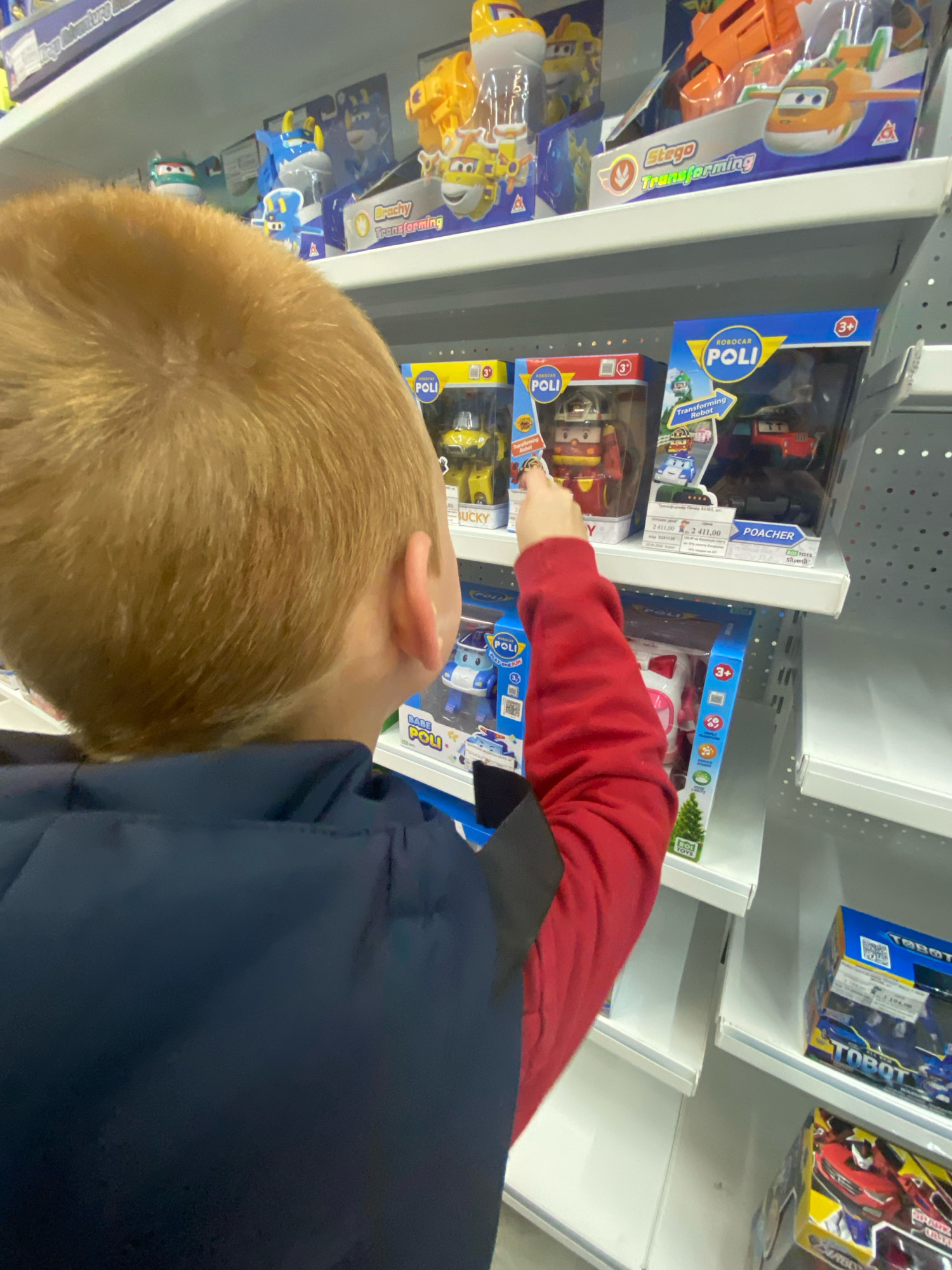 A young boy with blond hair and wearing a dark blue jacket and a red long-sleeved shirt is reaching for toys on a shelf in a store. The shelves are white and filled with various toy packages, primarily from the "Robocar Poli" and "Tobot" brands. Some of the toys appear to be transforming robots. The boy is positioned with his back to the camera, focusing on a specific toy package. The lighting suggests an indoor retail environment. The location is identified as a toy store in Chaikovsky, Russia.
