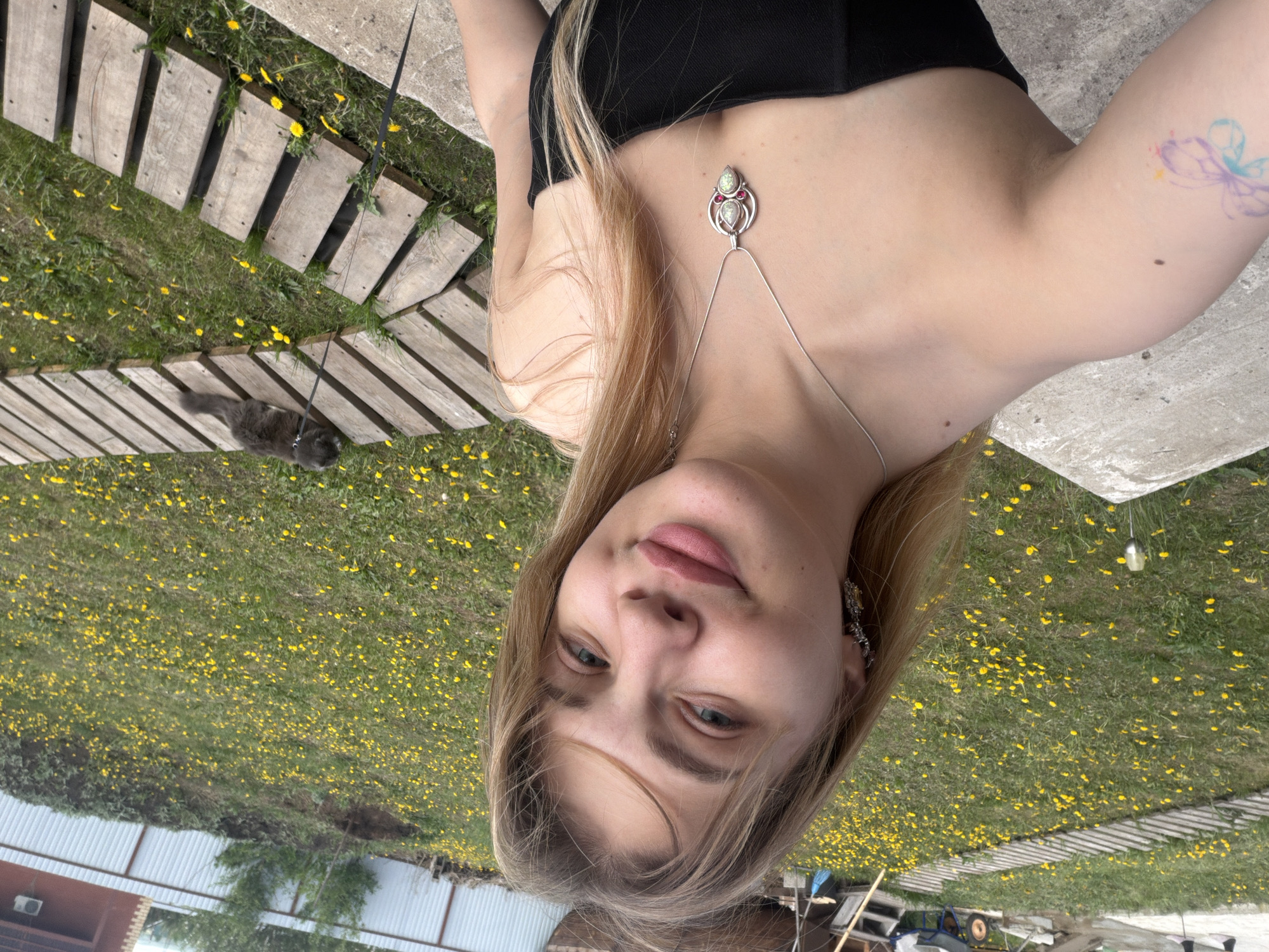A young woman poses for a selfie outdoors, lying on a concrete surface adjacent to a wooden walkway. The location appears to be a grassy area with numerous yellow dandelions in Большая Шильна, Russia. She is wearing a black strapless top and a silver body chain with a pendant. A gray cat is visible on the wooden walkway, attached to a leash. A tattoo is visible on the woman's upper arm. Behind her, a lawn area extends towards a partially visible building and various objects, possibly gardening tools or equipment.