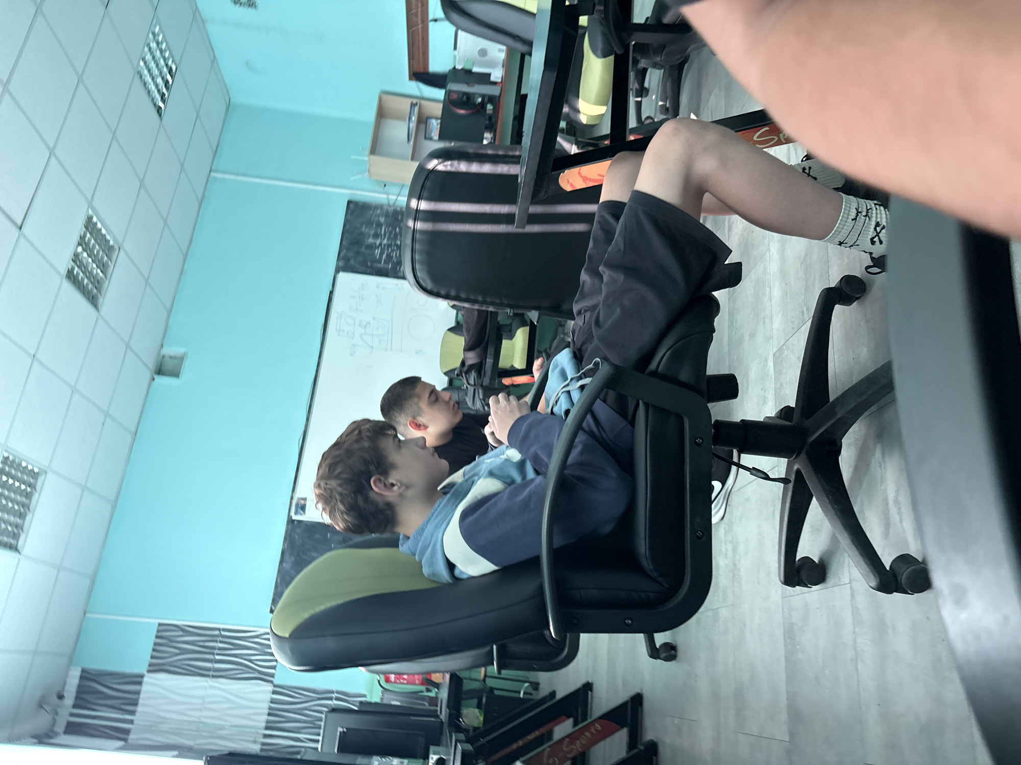 A young male with brown hair, wearing a blue and white hooded sweatshirt and black shorts, is seated in a black and green office chair. He is looking down at a mobile device in his hands. The chair is positioned in front of a desk, with other desks and chairs visible in the background. The room appears to be a computer lab or gaming center, with multiple computer monitors and gaming equipment. The walls are painted a light blue, and a whiteboard with writing is visible on the back wall. Fluorescent ceiling lights illuminate the space. A second young male, with short dark hair, is seated at a desk to the right, also looking at a device. The location is identified as Svoge, Bulgaria.