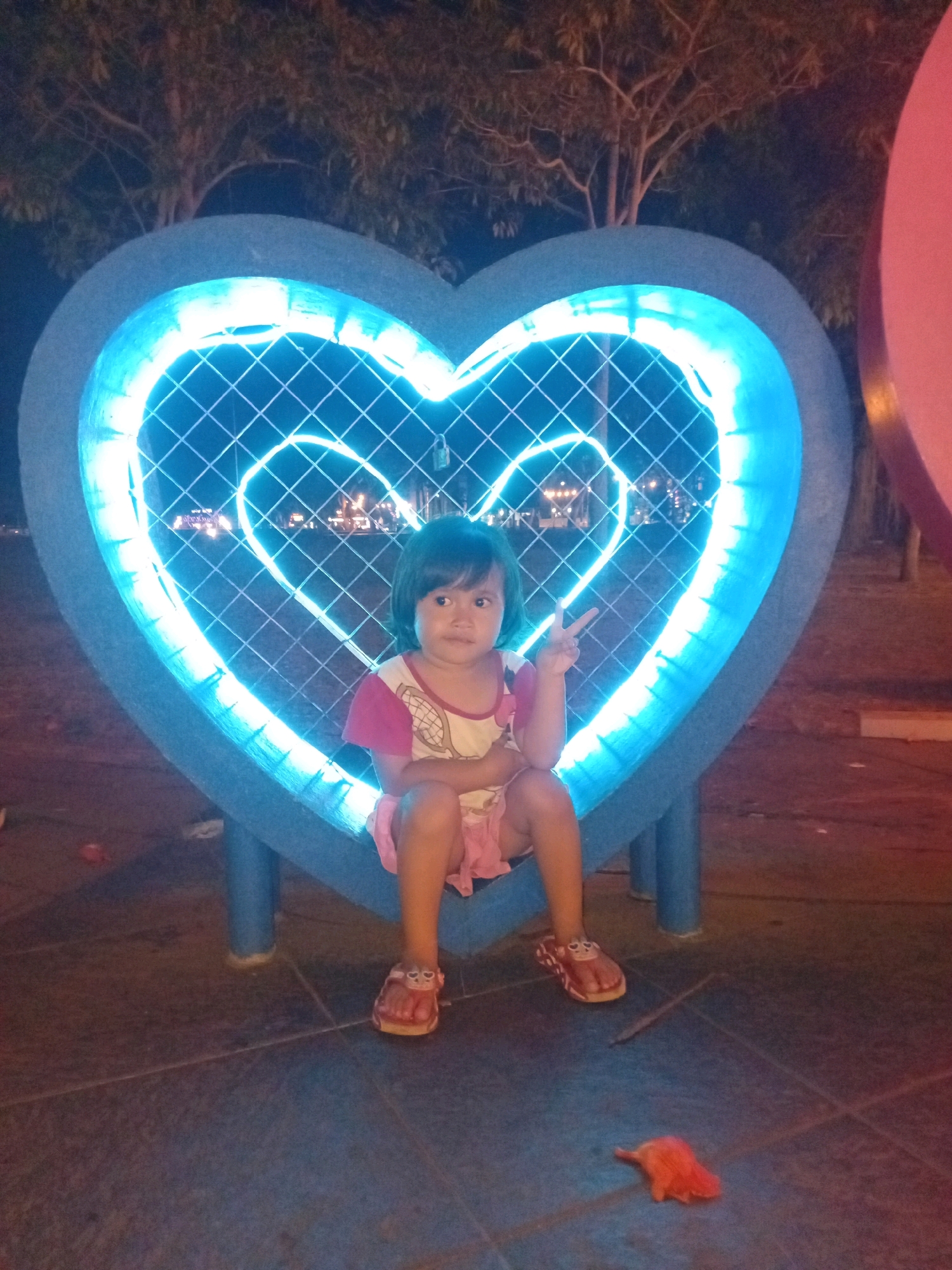 A young girl is sitting inside a large, blue, heart-shaped structure illuminated by blue neon lights. The heart structure has a diamond-patterned mesh inside. The girl is wearing a pink and white shirt and a pink skirt. She is barefoot with red sandals. She is making a peace sign with her right hand. The heart structure is on a paved surface. In the background, trees are visible. A pink object is on the ground to the right of the girl. The scene appears to be an outdoor public space at night. This image was taken in Purworejo, Indonesia.