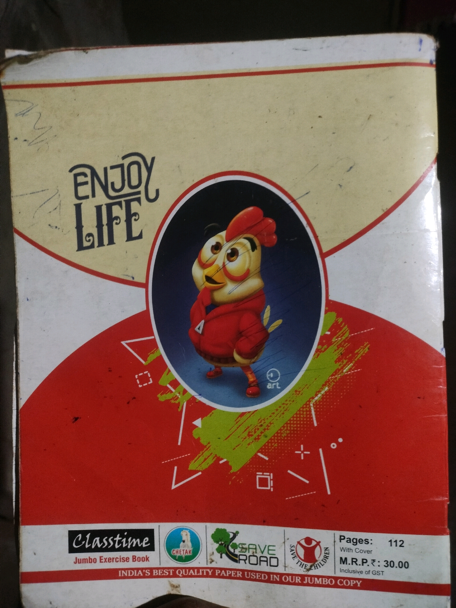This is a close-up, eye-level, indoor shot of a notebook. The notebook is predominantly red and yellow with a cartoon of a chicken wearing a red sweater in the center. Text on the notebook reads "ENJOY LIFE" in large, stylized letters, and "Classptime Jumbo Exercise Book" with the "Chetak" logo. Another section has text that reads "SAVE ROAD", "SAVE THE CHILDREN", and "Pages: 112 With Cover M.R.P.: 30.00 Inclusive of GST". The notebook is resting on an unseen surface. The lighting is dim and appears to be from above. There are some scratches and dust on the notebook, indicating it is used. No people are visible in the image. The location in the city of Raidandia, India is not depicted or suggested in this image.