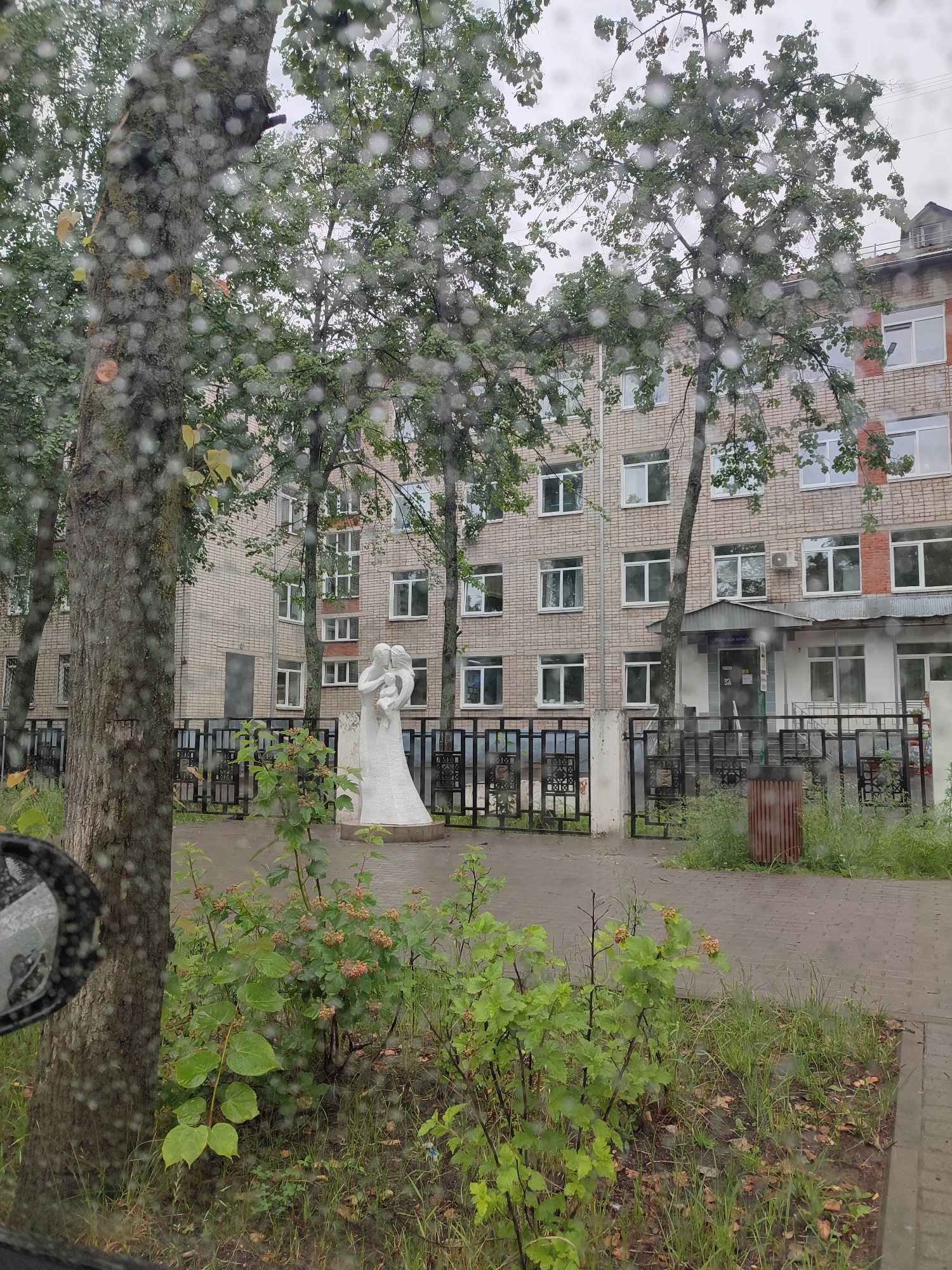 A white stone sculpture depicting a family, possibly parents embracing a child, stands in front of a light-brick building with multiple windows. The sculpture is situated on a paved area, behind a black metal fence. Green trees with lush foliage are visible on both sides of the scene, and in the foreground, there are bushes and grass. The ground appears wet, suggesting recent rain, and water droplets are visible on the camera lens, creating a blurred effect. The building is multi-storied, with a gray awning over an entrance. A trash can with a reddish-brown hue stands to the right of the fence. The overall impression is an outdoor scene in a somewhat overcast environment.