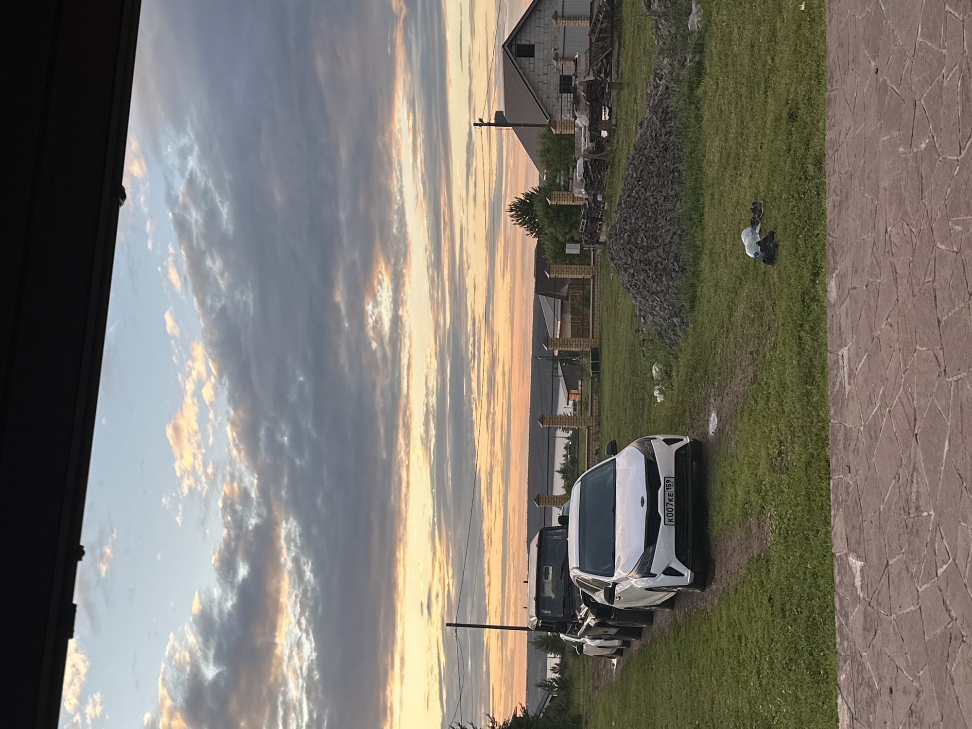 The image depicts a rural or semi-rural setting, likely in or near the village of Пепеляево, Russia, during a sunset. In the foreground, a paved patio area made of irregular, reddish-brown stones is visible. Behind this, a grassy yard contains a dirt path leading to a line of parked vehicles. The closest vehicle is a white Kia sedan with a license plate reading "KO07KE 159". Behind it are at least two dark-colored SUVs or off-road vehicles.
The midground shows a residential construction area. A large pile of dark gravel or crushed stone is prominent. Further back, a fence constructed of brick pillars and wire mesh separates the property from a building under construction, appearing to be made of concrete blocks with a pitched roof. Trees and other foliage are scattered throughout the landscape.
The background features a distant body of water, likely a lake or river, stretching across the horizon. The sky is dominated by a dramatic sunset with a mixture of scattered white and grey clouds illuminated by warm orange and yellow light. Power lines and a utility pole are visible against the sky, indicating the presence of infrastructure. No people are visible in the scene.