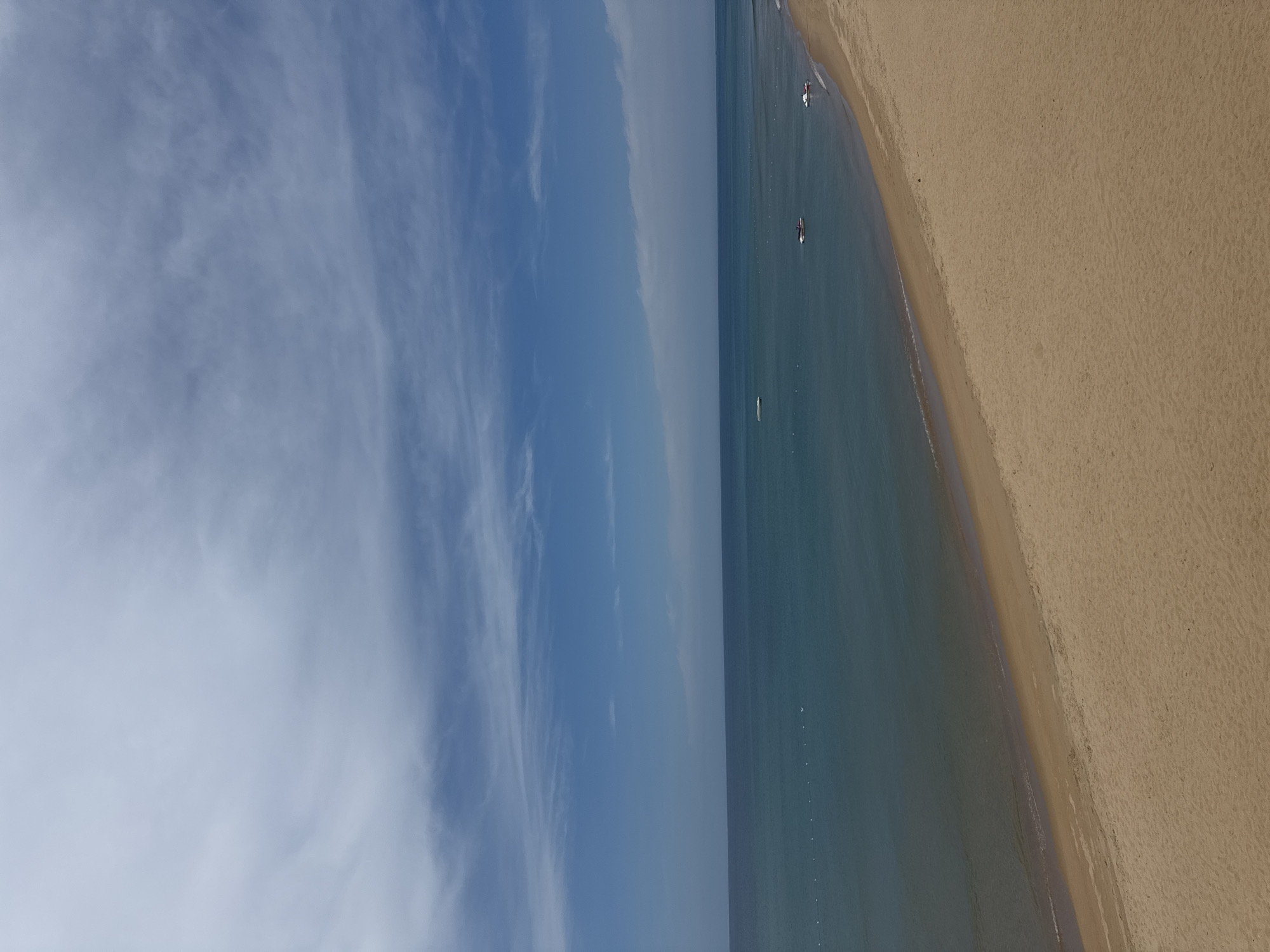 The image depicts a serene beach scene in Tuy Hoa, Vietnam, under a partially cloudy blue sky. The foreground is dominated by a vast expanse of light brown sand, showing subtle undulations and a few scattered dark specks, possibly debris or small stones. A gentle curve defines the edge of the beach, where it meets the calm, turquoise ocean.
The ocean stretches to the horizon, a gradient of deep blue in the distance transitioning to a lighter, more translucent shade closer to the shore. A faint, almost invisible line of small white buoys marks a boundary or demarcation in the water, running parallel to the coastline. Several small watercraft are visible in the sea. Closer to the shore, a white personal watercraft is positioned to the right, with a slightly darker object, possibly another watercraft or a small boat, to its left. Further out, a small white boat or floating object is visible near the center of the frame.
The sky above is a bright blue, streaked with wispy white clouds that create a delicate, feathery pattern across the upper half of the image. The overall impression is one of tranquility and natural beauty, characteristic of a coastal landscape.