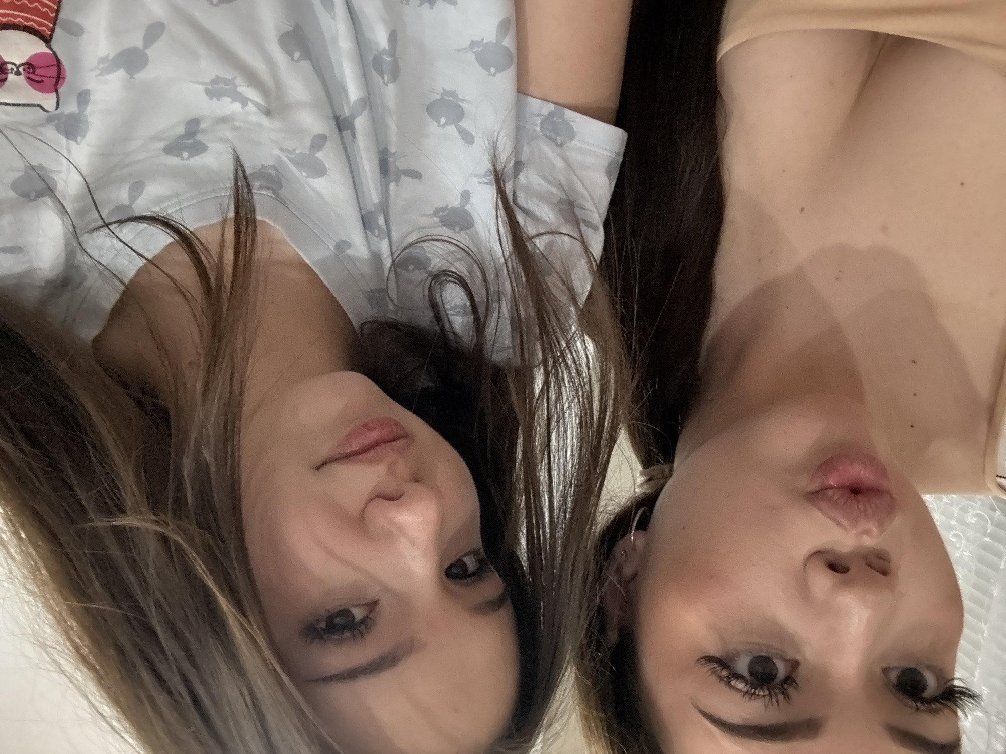 Two young women are featured in a close-up, intimate selfie, presented in a portrait orientation that has been rotated 90 degrees counter-clockwise, making them appear upside down. The setting is indoors, with a soft, out-of-focus background that suggests a casual home environment.
The woman on the left has long, dark brown hair that falls around her face. She is wearing a light-colored, possibly pajama-style top, featuring multiple small, grey, stylized cat-like figures that resemble Totoro characters. A larger character with a red and white striped bow is visible near her left shoulder. She looks directly at the camera with a gentle, slight smile, her expression appearing natural and serene.
The woman on the right is positioned next to her, also with long, dark brown hair. She is wearing a simple, light beige or nude-colored strapless top, which reveals a portion of her chest with a few moles. Her expression is playful, with her lips pursed into a "kiss face" or "duck face" and her eyes wide open. She has noticeable black eyeliner and long, dark eyelashes, possibly false, enhancing her gaze. Small silver hoop earrings are visible in her ears.
The image captures a casual and affectionate moment between the two, conveying a sense of familiarity or friendship. The lighting is soft and ambient, providing no specific time of day cues. The provided location context is Naberezhnye Chelny, Russia.
