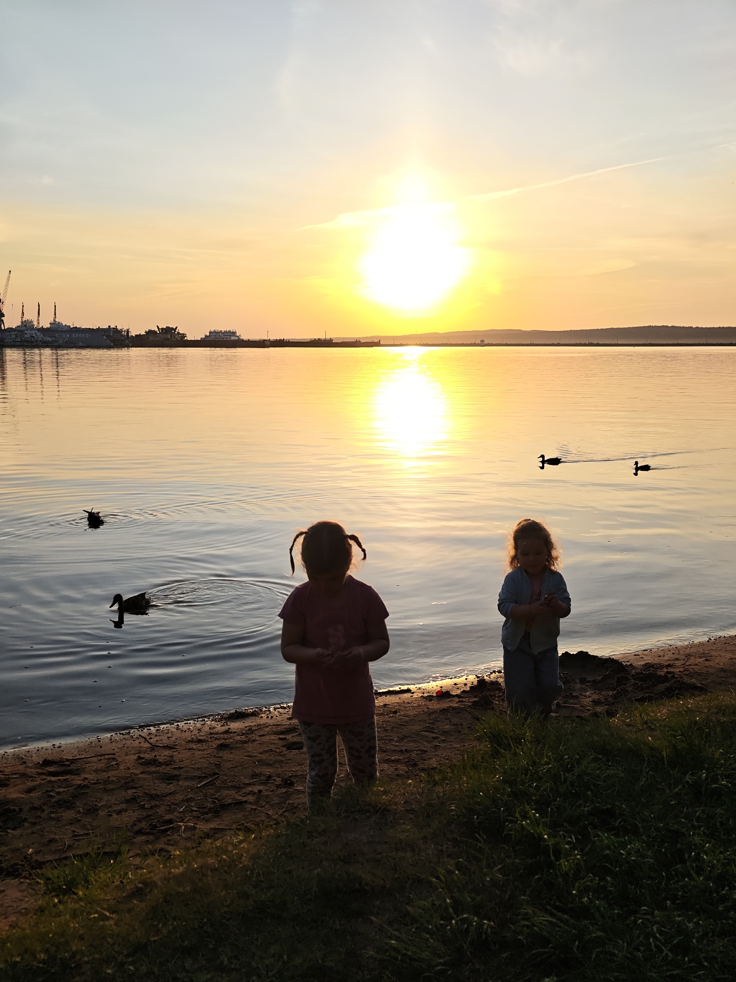 The image shows a sunset scene on the Kama River in Chaikovsky, Russia. Two young girls stand on the sandy beach. One with pigtails is wearing a dark shirt and patterned leggings. The other, with curly hair, wears a light-colored shirt and pants. In the water, several ducks are swimming. On the far side of the river, buildings and cranes of a port or industrial area are silhouetted against the setting sun. A contrail is visible in the sky.