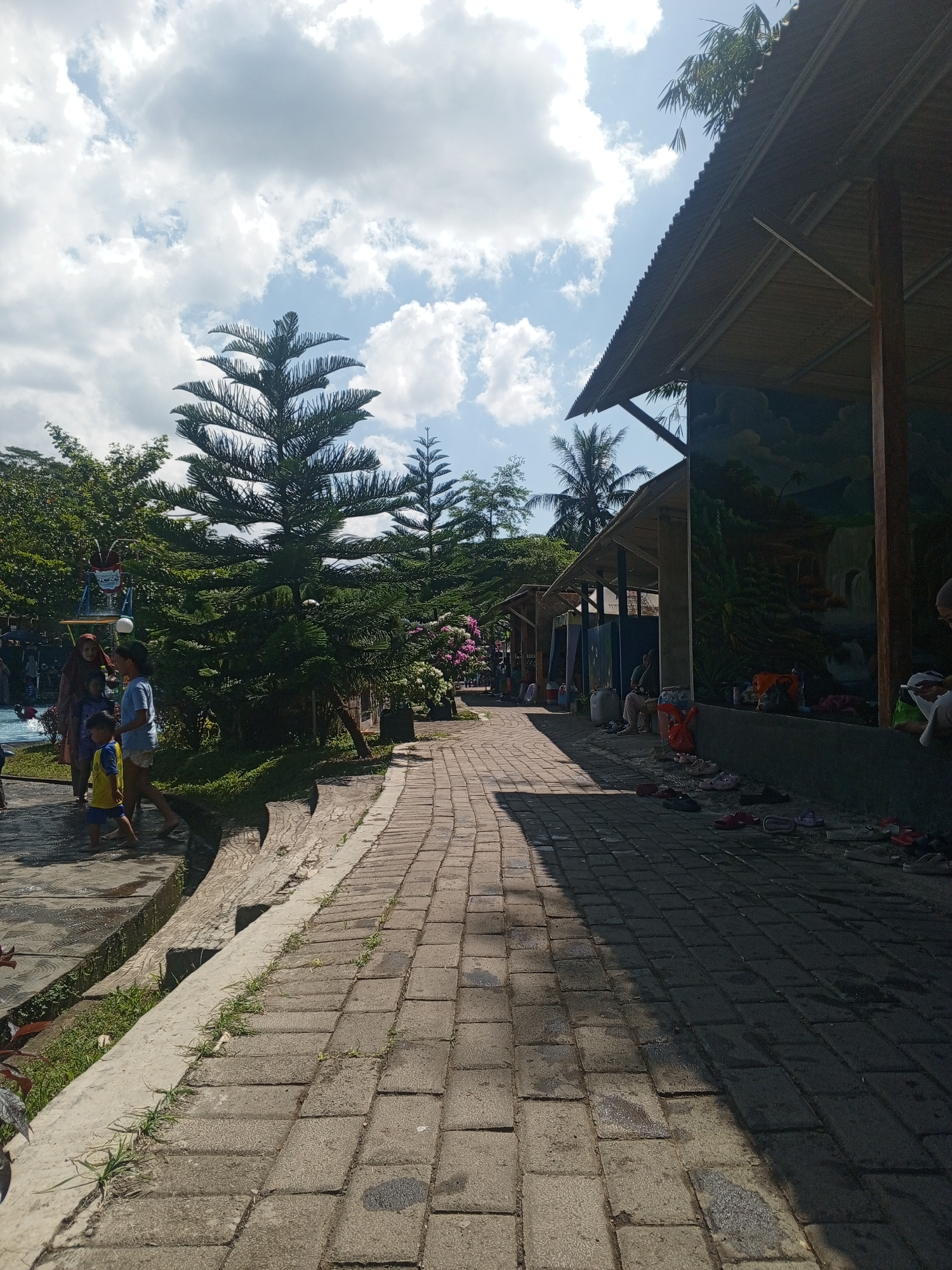 The image depicts a paved walkway at an outdoor establishment in Darsono, Indonesia. The walkway is constructed of rectangular stone pavers, with a slight gradient towards the right side of the frame. To the left of the walkway, there is a section with grass and what appears to be a shallow water feature, possibly a pool, with several people present, including adults and children. A tall, conical evergreen tree stands prominently in the mid-left of the image, behind the people. Beyond the tree, lush green foliage and other trees are visible.
To the right of the walkway, a covered structure runs along the length of the path. This structure features a corrugated metal roof supported by wooden posts. The walls of the structure appear to be decorated with a mural. Several people are seated or standing under this shelter, and a collection of footwear is visible on the ground in front of it. The environment suggests a leisure or recreational area, possibly a water park or a resort, given the presence of water and people in casual attire. The sky above is partly cloudy, with patches of blue sky visible. The lighting indicates it is daytime, with shadows cast by the structure and trees onto the walkway.