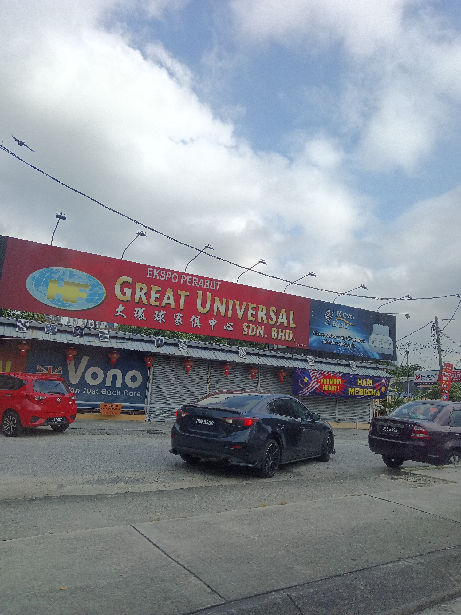 The image depicts a street scene in Kuala Lumpur, Malaysia, under a cloudy sky. A large red billboard prominently displays "EKSPOS PERABUT GREAT UNIVERSAL" with Chinese characters below, suggesting a furniture store. Adjacent to it is a smaller billboard advertising "KING KOIL" mattresses. Below the billboards, a row of shops is visible, some with closed shutters and decorative lanterns.
In the foreground, a dark-colored sedan with license plate VBM 5996 is parked on the road, facing away from the camera. To its left, a red hatchback with license plate RAA 7027 is also parked. Another dark-colored sedan, partially visible on the right, has the license plate JLS 5399.
A banner with the Malaysian flag and the words "PROMOSI HEBAT! HARI MERDEKA!" (Great Promotion! Independence Day!) is hung below the shopfronts. The environment appears to be an urban commercial area with power lines and streetlights present. No people are clearly visible in the image.