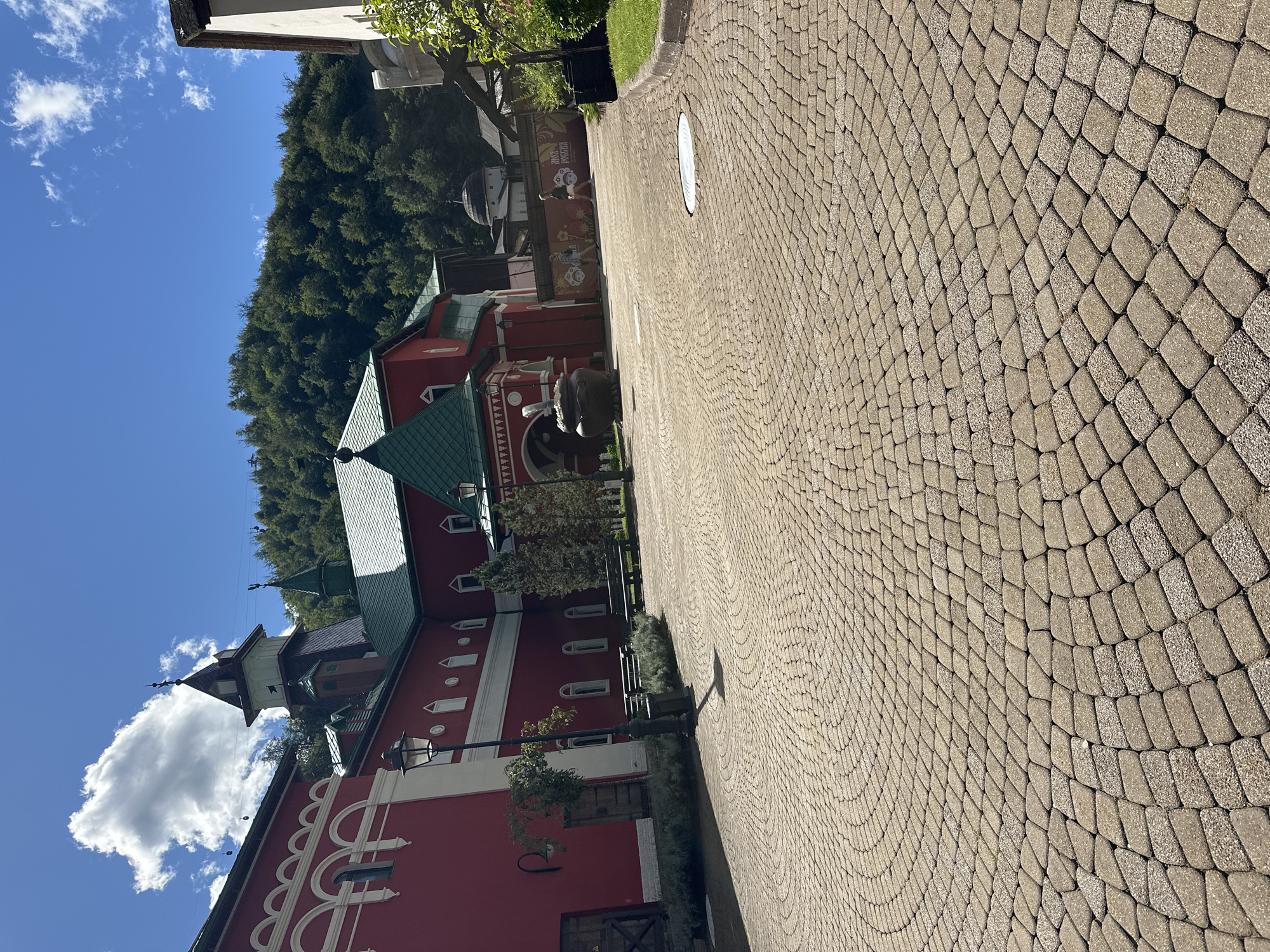 This is a photograph taken on a sunny day in Estosadok, Russia. The foreground is dominated by a circular pattern of cobblestones. To the left, a bright red building with white decorative trim and arched windows is visible. A black lamppost stands in front of this building. In the background, a large red building with a green roof and a tower is prominent. The building has smaller towers and intricate architectural details. Behind the buildings, a lush green forest covers a hillside under a blue sky with scattered clouds. In the middle ground, two people are walking away from the camera on the cobblestone path.