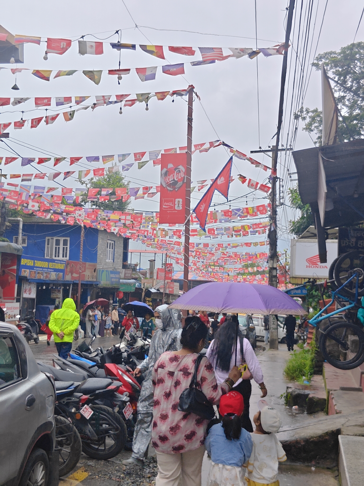 In Pokhara, Nepal, a street scene unfolds under a cloudy sky, with strings of international flags draped overhead. The flags, representing various countries, create a festive yet somewhat subdued atmosphere, possibly due to the overcast weather.
In the foreground, a person in a bright yellow raincoat stands facing away from the camera, with their back to the viewer. To their right, several motorcycles are parked, their surfaces glistening from recent rain.
Further back, a group of people walks along a narrow path, some holding umbrellas to shield themselves from the rain. A woman in a patterned shirt is in the center, holding a yellow camera. Two children, one in a red cap and the other in a white cap, are walking in front of her.
Buildings line the street, with signs indicating businesses such as "BIPISHA TANDOORI RESTAURANT." Coca-Cola branding is visible on a large red banner hanging overhead. The presence of power lines and poles suggests an urban setting. The overall scene depicts a typical day in a Nepalese city, with the added visual element of numerous national flags celebrating international connection.