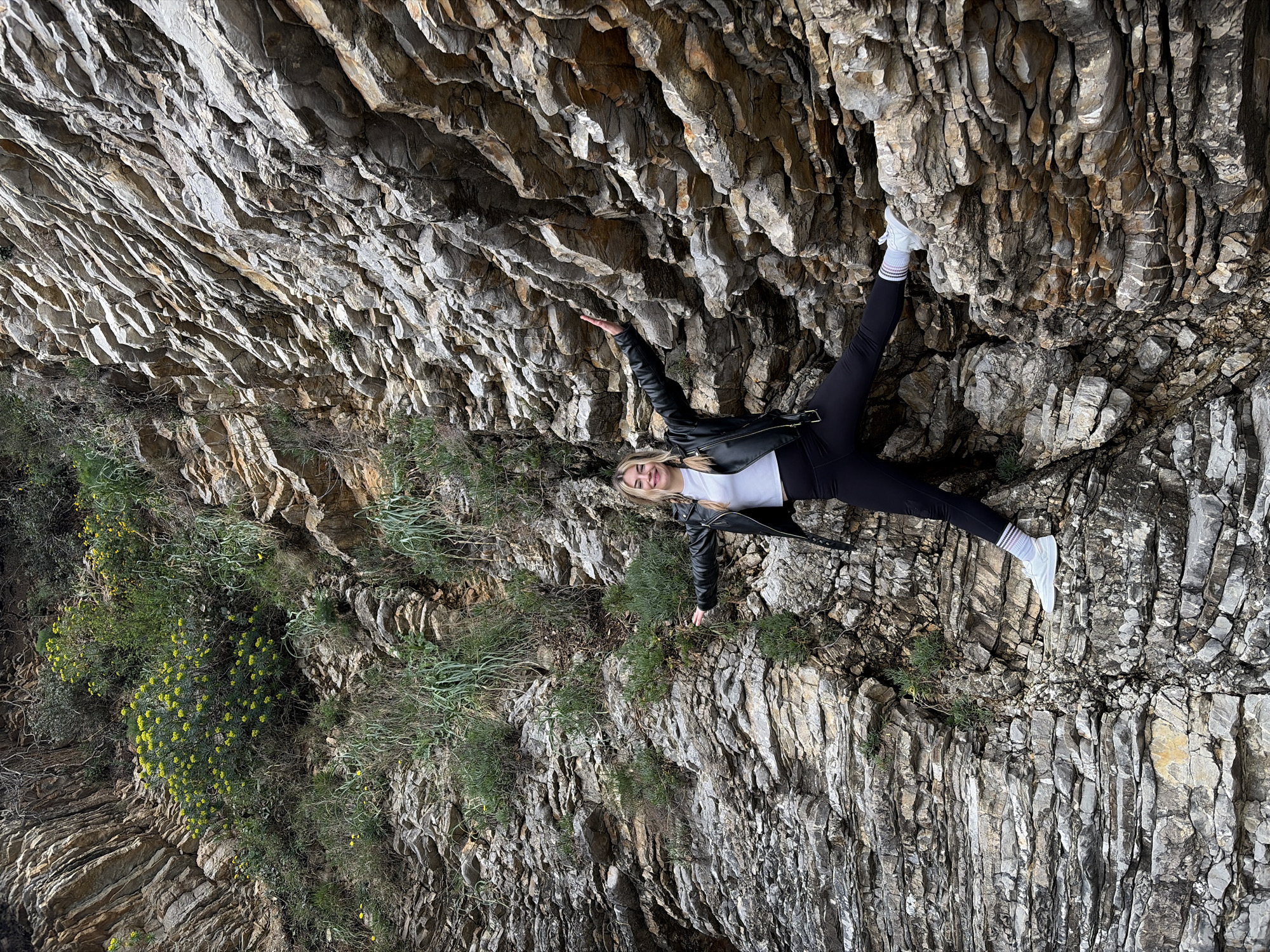 A photograph depicts a person posed against a cliff face. The person, a young female with blonde hair, is wearing a black leather jacket over a white shirt, black leggings, and white sneakers with striped socks. She is positioned with one arm outstretched and the other leg extended, creating a dynamic pose. The cliff face is composed of layered rock formations in shades of grey and brown, with some vegetation growing along the base. The location appears to be a natural environment, possibly along a coastline or hillside near Budva, Montenegro, given the style of rock formations and plants.