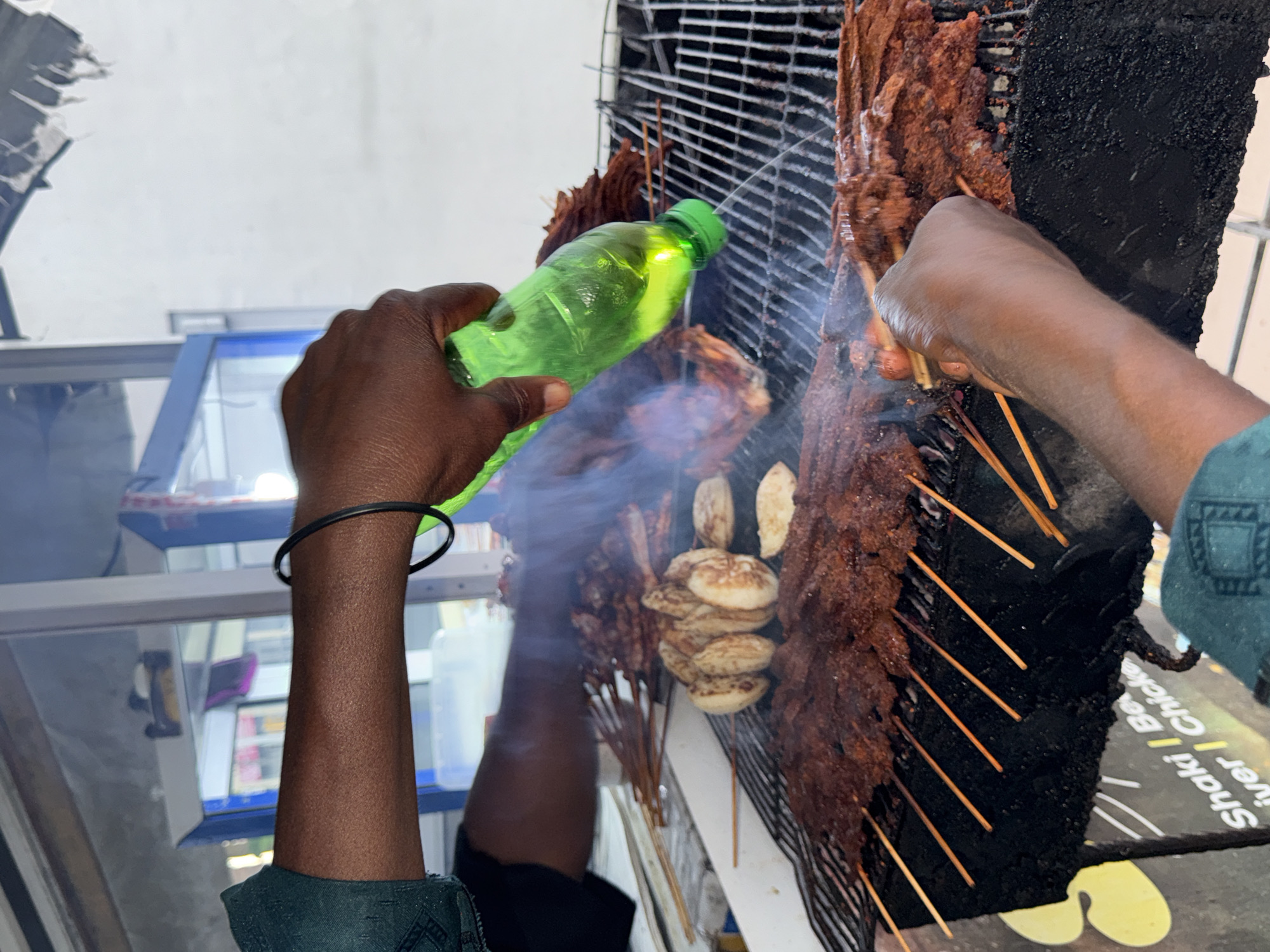 In Ado, Nigeria, a street vendor is grilling skewers of marinated meat. The scene is captured from a close-up, eye-level perspective, focusing on the hands of the vendor. The vendor, with dark skin and wearing a green top, is pouring a clear, green liquid from a plastic bottle onto the grill. The liquid is creating steam and smoke as it hits the hot grill and the meat.
The grill itself is made of metal grates and is covered with numerous skewers of raw, marinated meat, appearing to be beef or goat. There are also some round, flat items on the grill, possibly plantains or yams, also being grilled. The meat skewers are arranged in dense rows, with wooden skewers protruding upwards.
The background shows a white wall and a portion of what appears to be a shop or stall with blue and white accents. Some indistinct items are visible through a glass panel in the background. The overall impression is a busy, outdoor food stall in a Nigerian city, highlighting the process of grilling street food. The image emphasizes the sensory experience of cooking, with the visible steam and the implied heat from the grill.