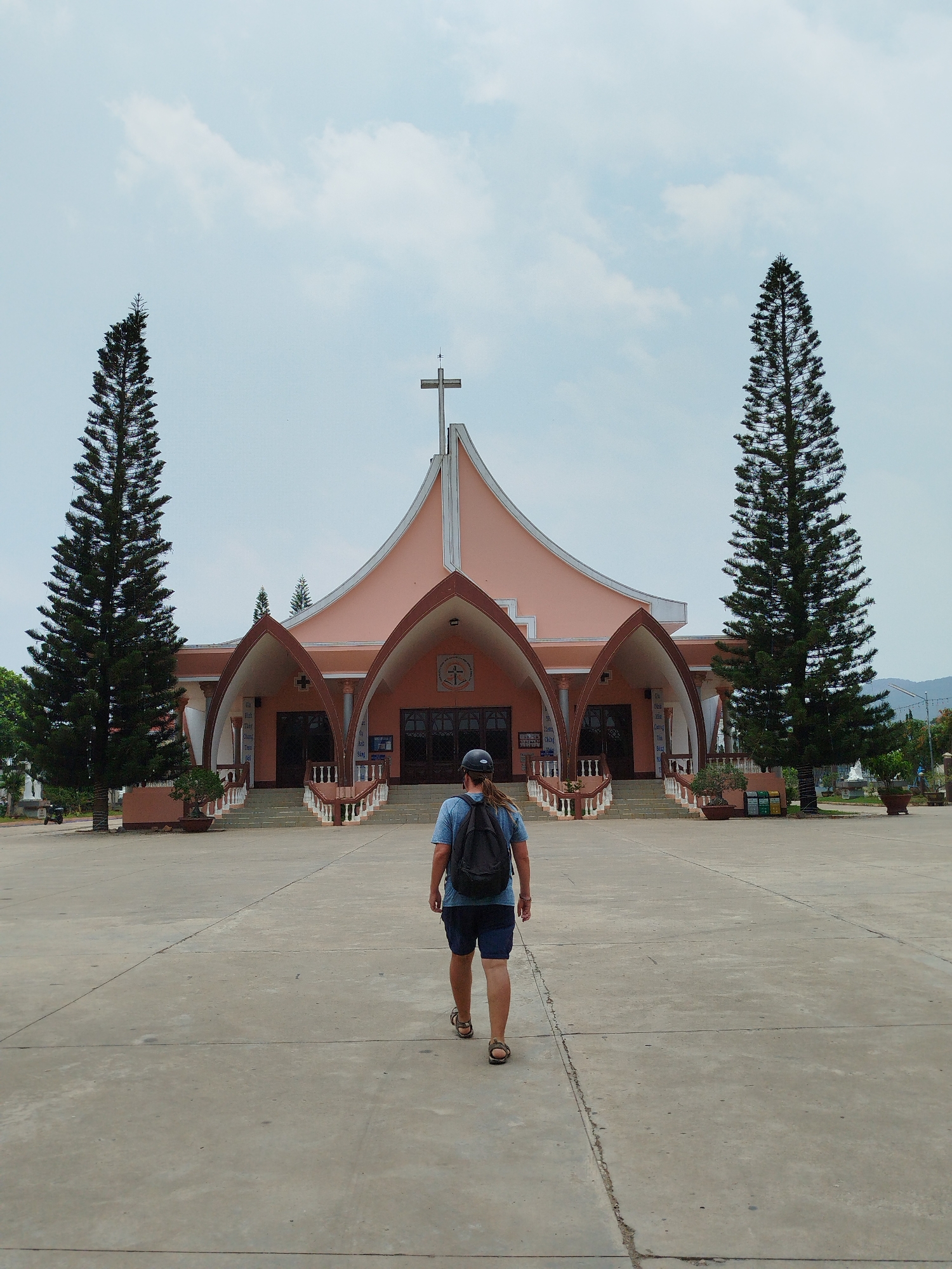 A person with a backpack walks toward a church with a pink façade in Phú Thạnh, Vietnam. The church has a cross on its roof and two tall trees flanking the entrance. The foreground is a concrete courtyard. The sky is overcast.