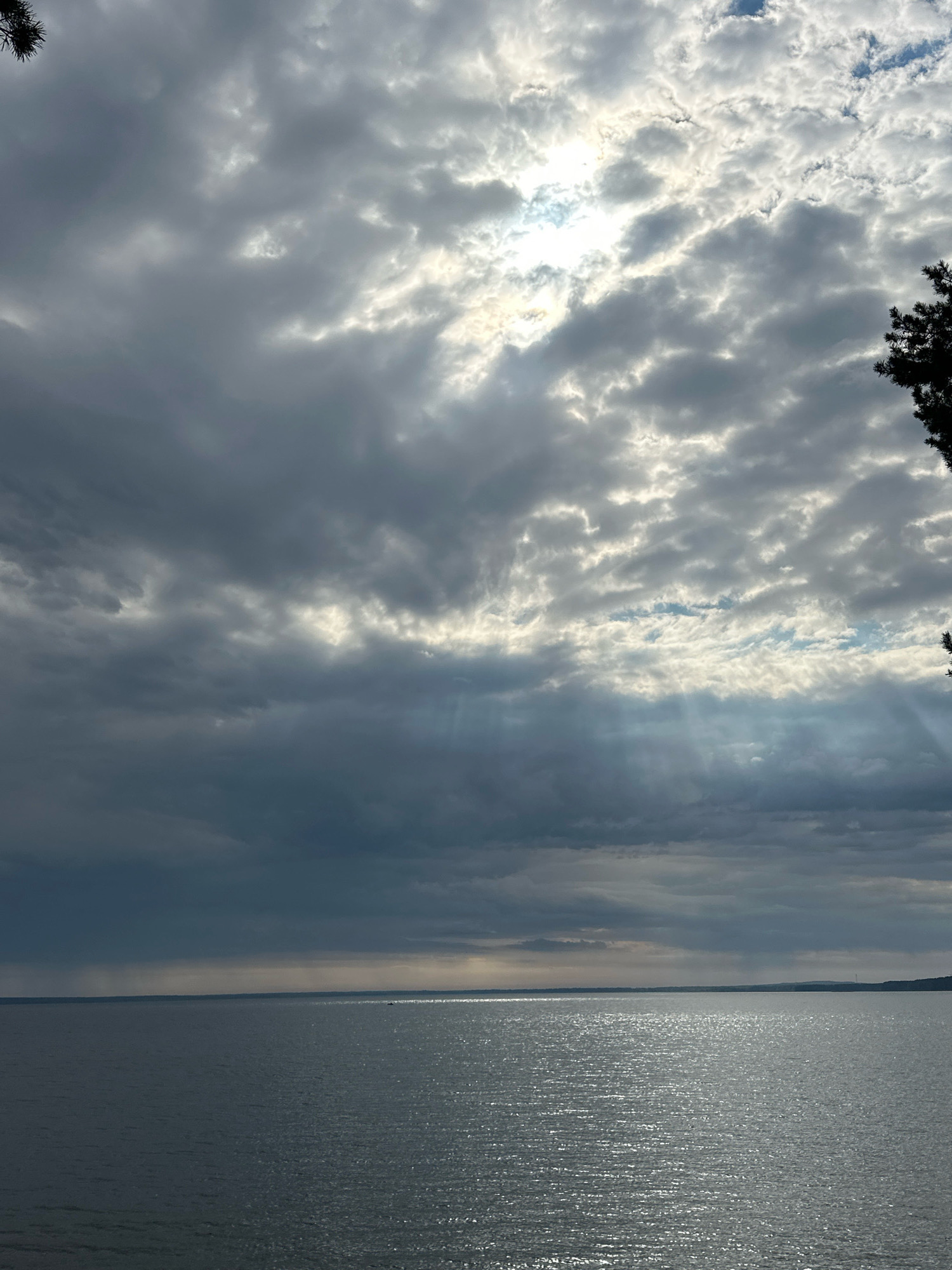 The image shows a wide expanse of water under a cloudy sky. Sunbeams break through the clouds, creating streaks of light that illuminate the water's surface. The water appears calm with ripples reflecting the light. In the distance, a faint shoreline with some vegetation is visible. A dark silhouette of a tree branch extends from the upper right corner of the frame. The location is identified as Нарацкі сельскі Савет, Belarus.