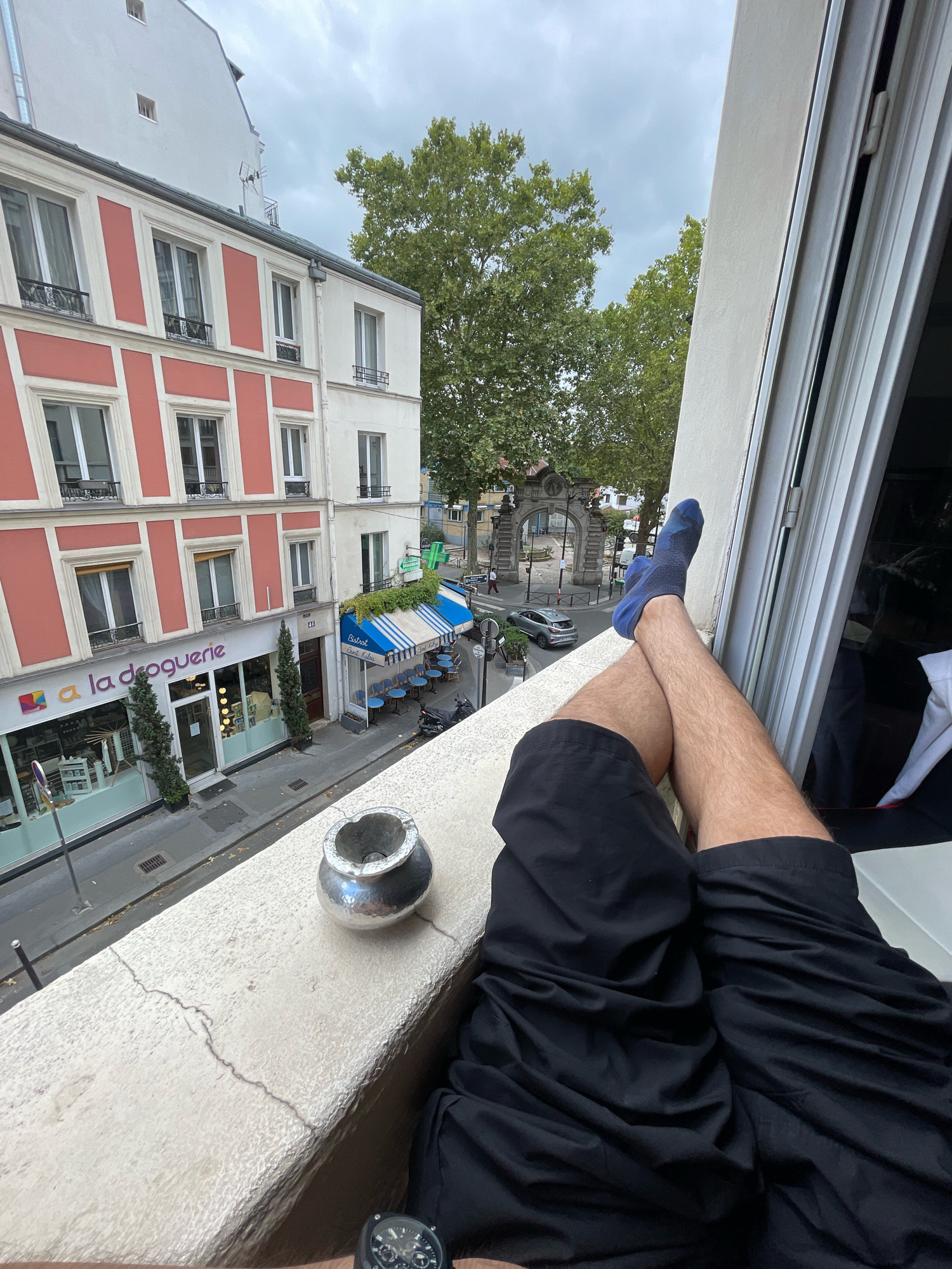 A person's legs are propped up on a concrete ledge overlooking a street in Paris, France. The individual is wearing black shorts and blue socks. A silver ashtray rests on the ledge to the left of the legs. In the background, there is a building with red and white facades, an outdoor cafe with blue umbrellas and chairs, and a stone archway. The sky is overcast. The street below has a few parked cars and a scooter. The location is identifiable as Paris due to the architectural style of the buildings, the presence of a cafe, and the general urban setting typical of the city. The stone archway in the distance also appears to be a Parisian landmark, possibly the Porte Saint-Denis.