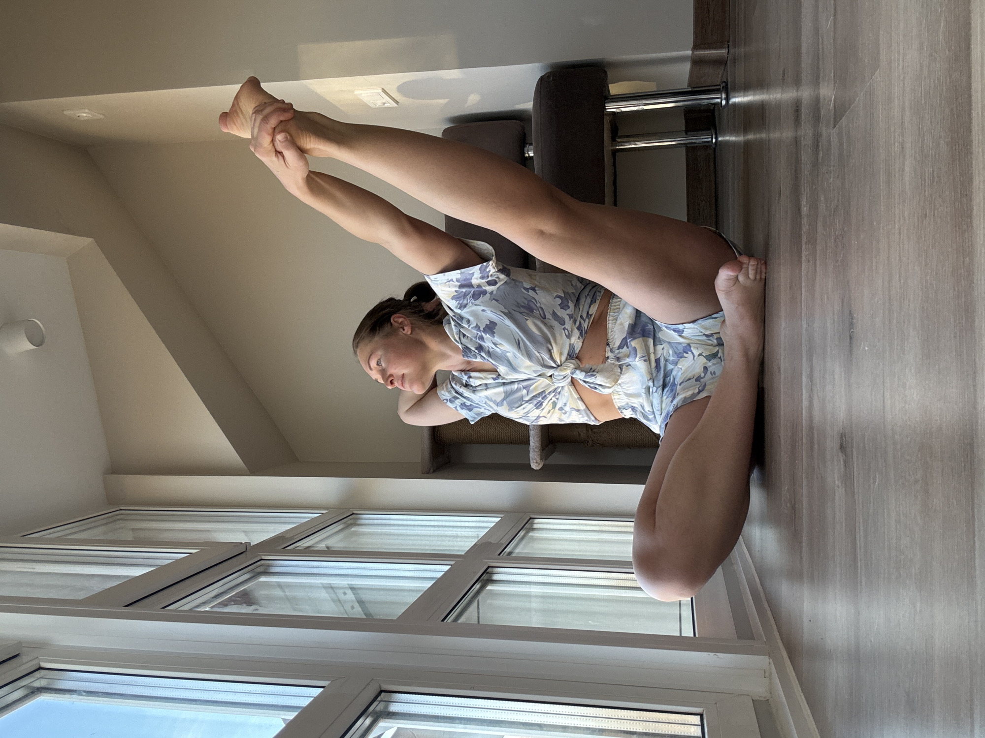 A woman is performing a yoga pose indoors. She is kneeling with one leg bent behind her and the other extended upward, holding her feet with her hands. She is wearing a floral patterned top and shorts. The room has a large window with a view of the sky and possibly other buildings, a light-colored wooden floor, and a piece of furniture with metal legs. The photo appears to have been taken in a residential setting. The location cannot be confirmed as the city of Chaikovsky, Russia.