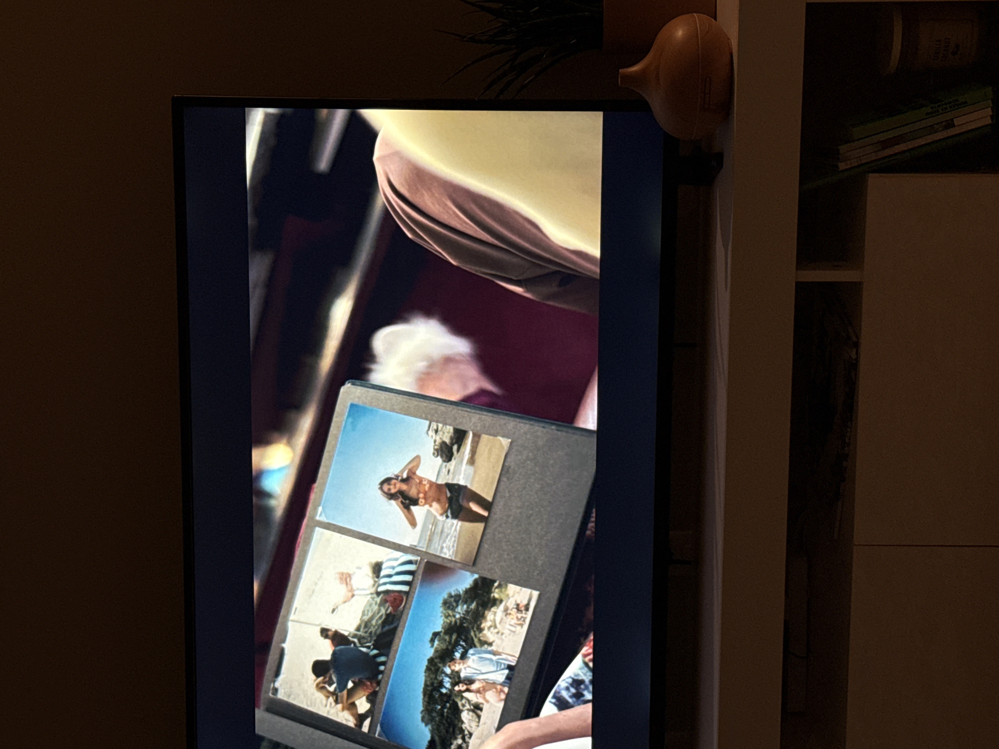 A television screen displays a photograph album with several photos of people on a beach. One photo shows a woman in a bikini posing on the sand. Another photo shows a couple standing near trees, and a third photo shows a group of people relaxing on the beach. The television is placed on a white shelf unit, and a brown diffuser is visible on the shelf to the right of the television. The background is dark, suggesting the room is dimly lit. A plant in a brown pot is also visible on the shelf. The location is identified as Greenville, United States of America.