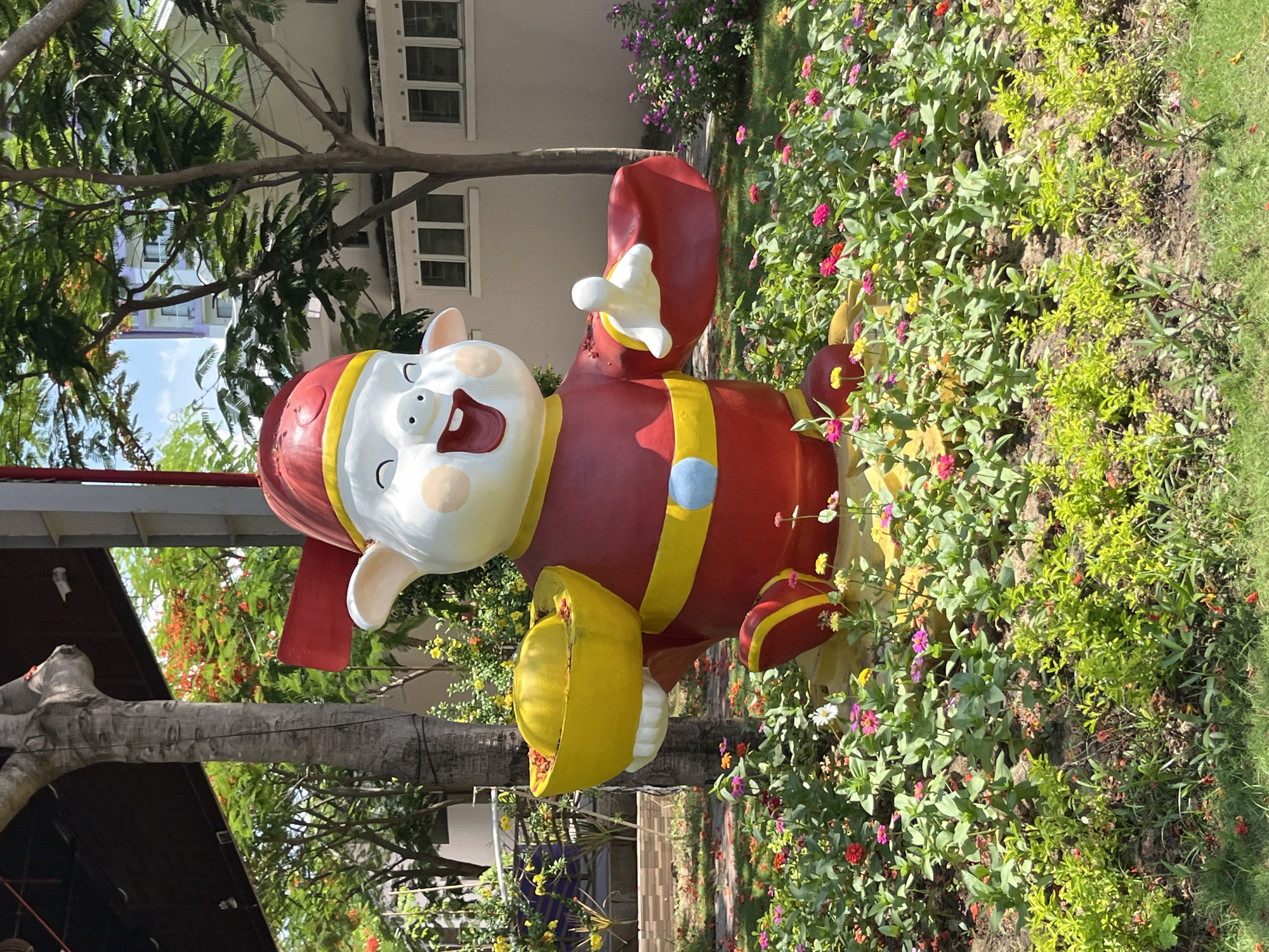 A large, colorful statue of a pig wearing a red hat and robe, holding a golden bowl filled with golden nuggets, is positioned in a garden filled with small, vibrant flowers. The statue appears to be a festive decoration, possibly for a holiday. Behind the statue, a building with multiple windows is visible, and trees with green leaves and some red blossoms are scattered throughout the scene. The overall impression is one of a well-maintained outdoor space, likely in a city, given the presence of the building and the manicured garden. The statue's design and the gold nuggets suggest a theme of prosperity and good fortune. The location is identified as Dong Ninh Hoa Ward, Vietnam.