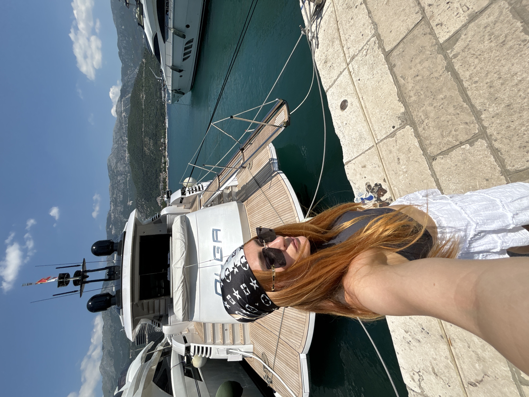 A woman with long, reddish-orange hair, wearing a black and white patterned bandana, sunglasses, a black top, and a white skirt, stands on a paved stone walkway by a marina. She is holding a phone to take a selfie. Behind her, a large white yacht with the name "OLGA" visible on its stern is docked in the turquoise water. The yacht has a wooden deck and a retractable gangway. Other yachts and a coastline with lush green mountains and buildings are visible in the background under a clear blue sky with scattered clouds. The scene is located in Budva, Montenegro, likely near the marina.
