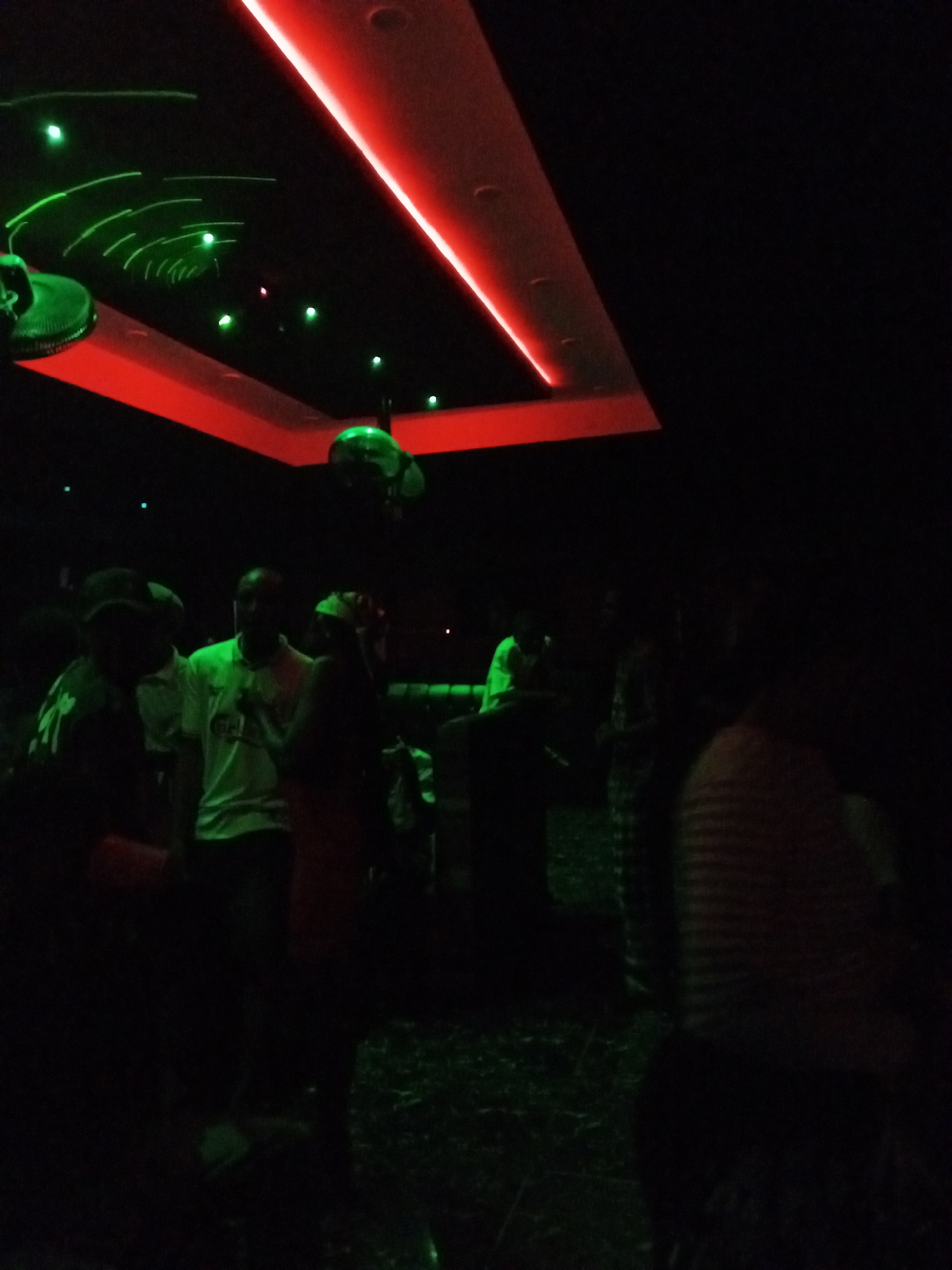 The image depicts the interior of a dimly lit establishment in Lokoja, Nigeria. The ceiling has recessed lighting with a red band and green laser lights. Several people are present, but their features are obscured by the low light conditions. One individual is wearing a light-colored shirt, while others have indistinct clothing. The environment suggests a nighttime social gathering, potentially a bar or club. The flooring appears patterned.