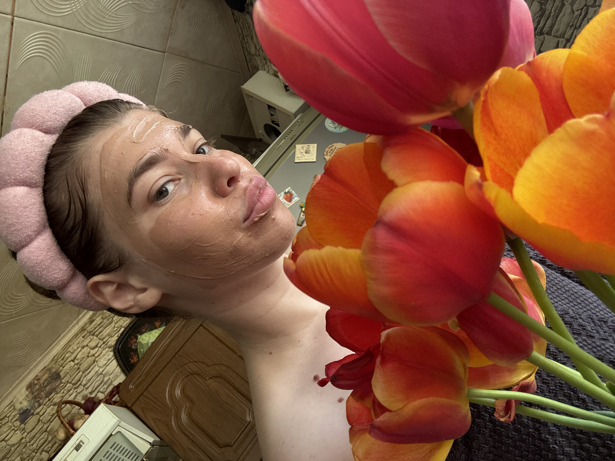 A woman wearing a pink headband and a facial mask is the primary subject of this selfie-style photograph. She is positioned to the left side of the frame and is angled towards the camera. To the right, a bouquet of tulips in shades of red, orange, and yellow partially obscures the view.
In the background, a kitchen setting is visible. A kitchen cabinet and a refrigerator are present, along with some unidentified objects placed on top of the refrigerator. The overall lighting suggests an indoor environment. The location is not Мещёры, Russia, because there is no evidence of geographic location in the photo.