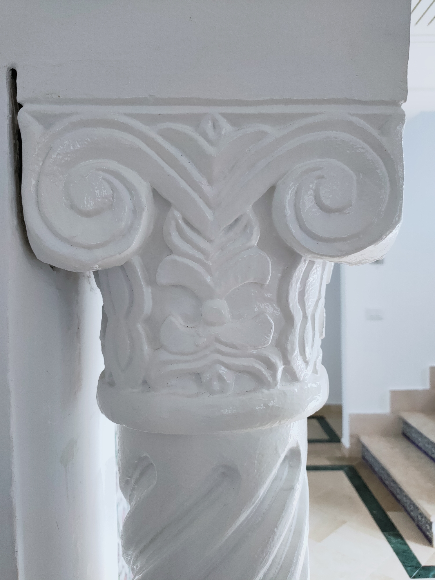 The image is a close-up, eye-level, outdoor shot of a white, ornate column with carved details. The column is the primary subject, taking up most of the frame. The top of the column features decorative volutes and a floral motif. Below this, the shaft of the column has a twisted, fluted design. The column appears to be part of an architectural feature, possibly an entrance or pathway, as indicated by the blurred background which shows a tiled floor with green and cream geometric patterns and a staircase with blue and white patterned risers. The lighting is natural and bright, suggesting daytime. The location is identified as Mrezga, Tunisia. There are no people visible in the image.