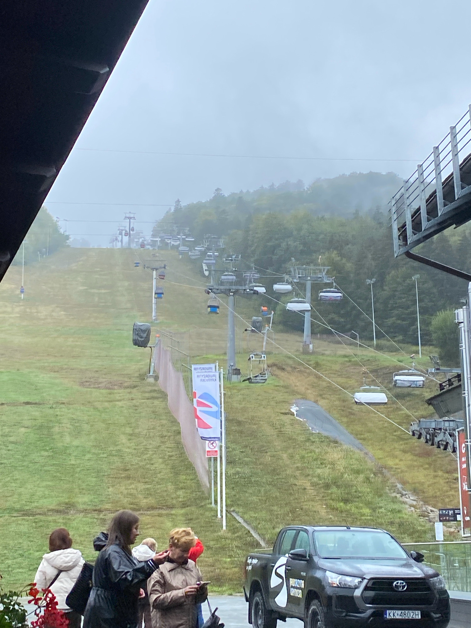 The image depicts a ski resort in Krynica-Zdrój, Poland, on a hazy, overcast day. In the foreground, several people are standing near a black Toyota pickup truck. The truck has a white logo on its side and a license plate reading "KK 480ZH". Behind the people, a grassy slope stretches upwards, dotted with numerous ski lifts and gondolas, indicating the resort's operational status. The ski slope is partially covered in grass, with some bare patches. A tall, modern ski lift structure is visible on the right side of the image. The background is dominated by a misty forest-covered hillside, further emphasizing the atmospheric conditions. Two large banners with Polish text and abstract graphics are displayed near the base of the slope. The overall impression is of a serene, perhaps slightly melancholic, mountain landscape with a functioning ski resort.