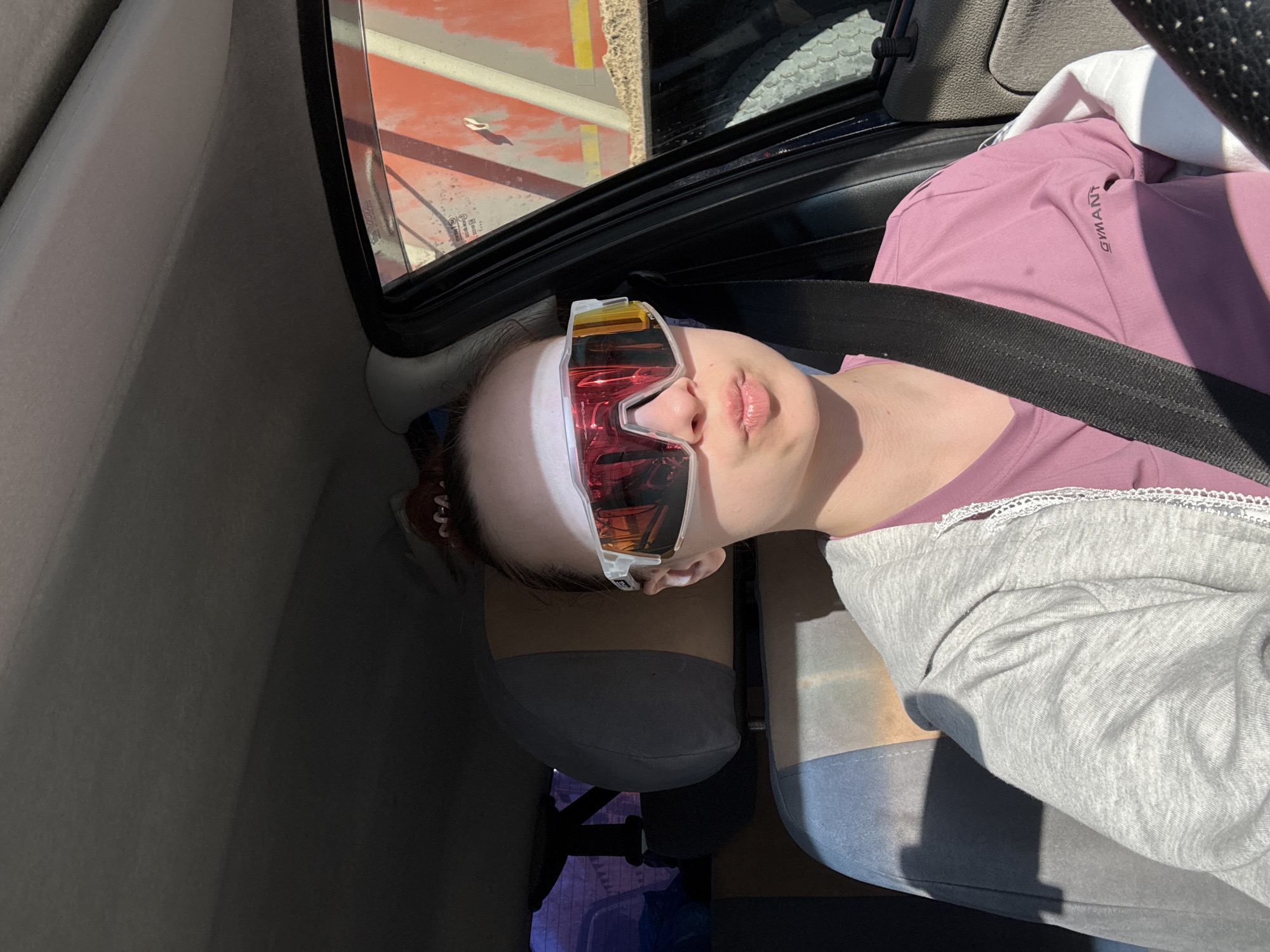 A woman is lying in the passenger seat of a car, wearing a seatbelt and large sunglasses. She is wearing a purple shirt with the word "SIMANT" printed on it and a grey jacket. A white headband covers her forehead. The car's interior is visible. Through the window, a glimpse of an urban landscape with red and beige structures is seen. Without more context, it is not possible to confirm that the location is Chaikovsky, Russia.