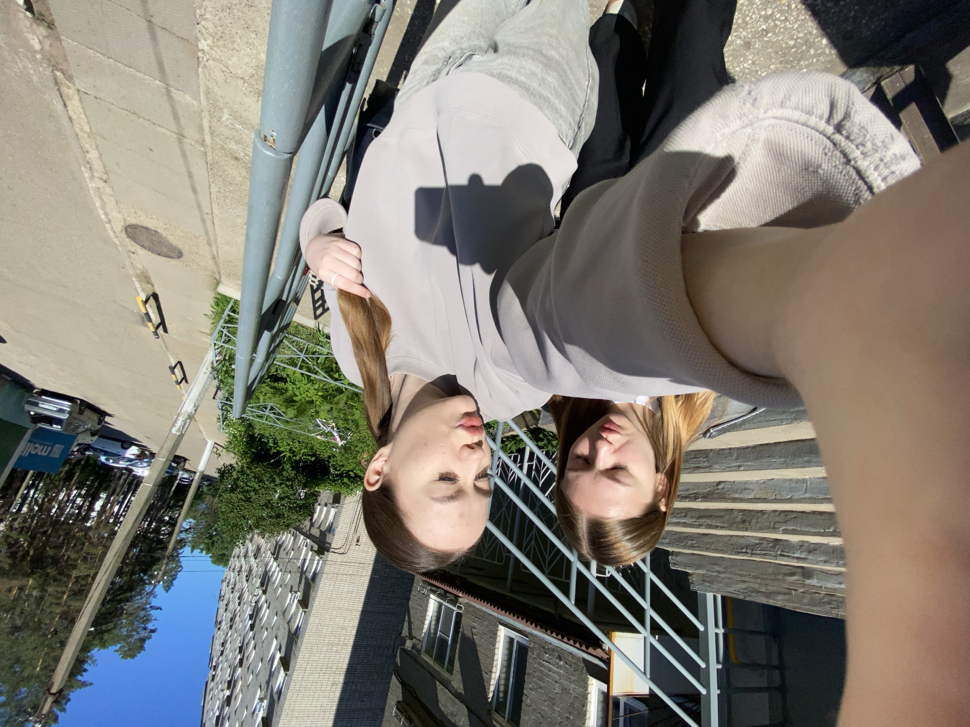 Two young women are taking a selfie outdoors on a sunny day. The woman in the foreground is in focus, wearing a light grey sweatshirt and light grey pants, with her hair pulled back in a ponytail. She is puckering her lips for the photo and touching her hair. The second woman, slightly out of focus, is standing next to her and looking down with her lips also pursed. They are positioned on outdoor stairs with a metal railing. In the background, a multi-story apartment building and tall pine trees are visible, suggesting a residential area in the city of Chaikovsky, Russia. A sidewalk with a manhole cover and parked cars is also present.
