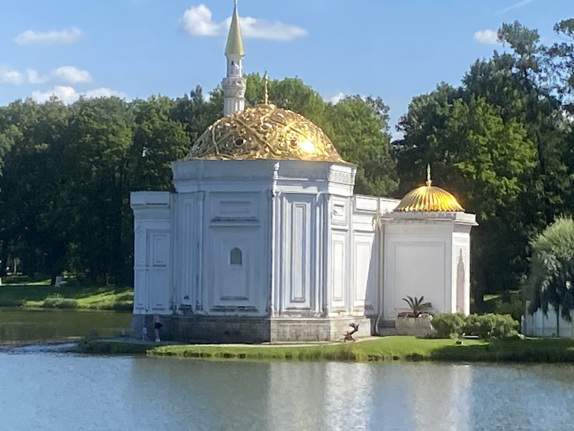 An exterior, eye-level, wide shot captures a white, classical-style building with a golden dome and minaret on the shore of a body of water. The building is the Turkish Bath, part of the Catherine Palace complex in Pushkin, Russia. The building has white walls with decorative vertical fluting and arched windows. The main dome is large and elaborately decorated in gold, with a smaller golden dome on a connected structure. A slender, golden minaret rises from behind the main dome. Green trees form a dense backdrop behind the building, and the sky above is a clear blue with a few scattered white clouds. The water in the foreground reflects the sky and the building. Two small figures, one in black shorts and a white top, and another in a blue top and black shorts, are visible standing on the edge of the building's foundation, near the water. Another small, dark figure is partially visible on the grass near the water's edge to the right of the building. The grass is green and well-maintained, with some small bushes and a potted plant.