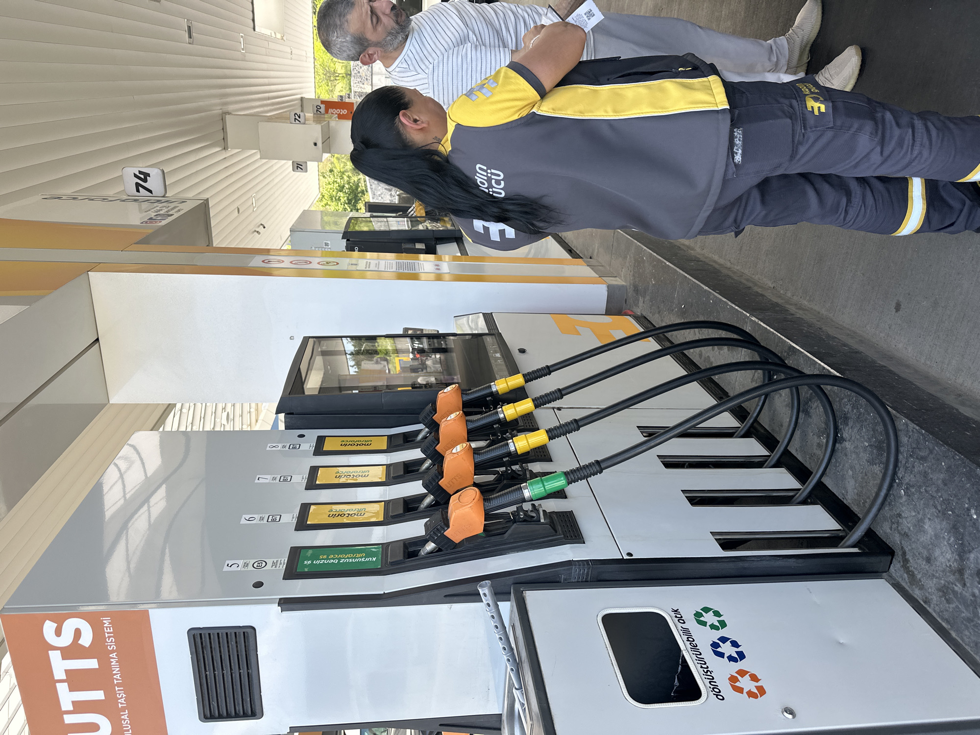 The scene depicts a gas station, likely in Gaziosmanpaşa, Turkey, based on the Turkish text and the style of the gas pump. A woman wearing a uniform is interacting with a customer near a gas pump. The pump has a display showing numbers 5 through 8, and nozzles indicating different fuel types. A recycling receptacle is visible on the pump. The station has a covered roof, and daylight suggests an outdoor setting.