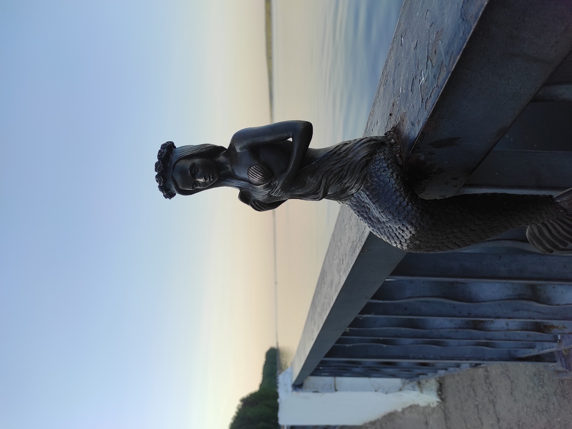 The image depicts a dark, metallic statue of a mermaid perched on a grey concrete and steel structure. The statue is positioned so that the mermaid appears to be looking out over a body of water, likely a lake or river, towards the horizon. The sky is a gradient of blue at the top transitioning to a pale yellow near the horizon, suggesting either dawn or dusk.
The statue has visible details such as scales on the tail, a seashell motif on the chest, and a floral wreath on the head. The steel structure appears to be part of a bridge or pier, with multiple support beams visible beneath the concrete surface on which the statue rests. A small amount of greenery is visible in the distance.
The location is provided as Unknown, Unknown.