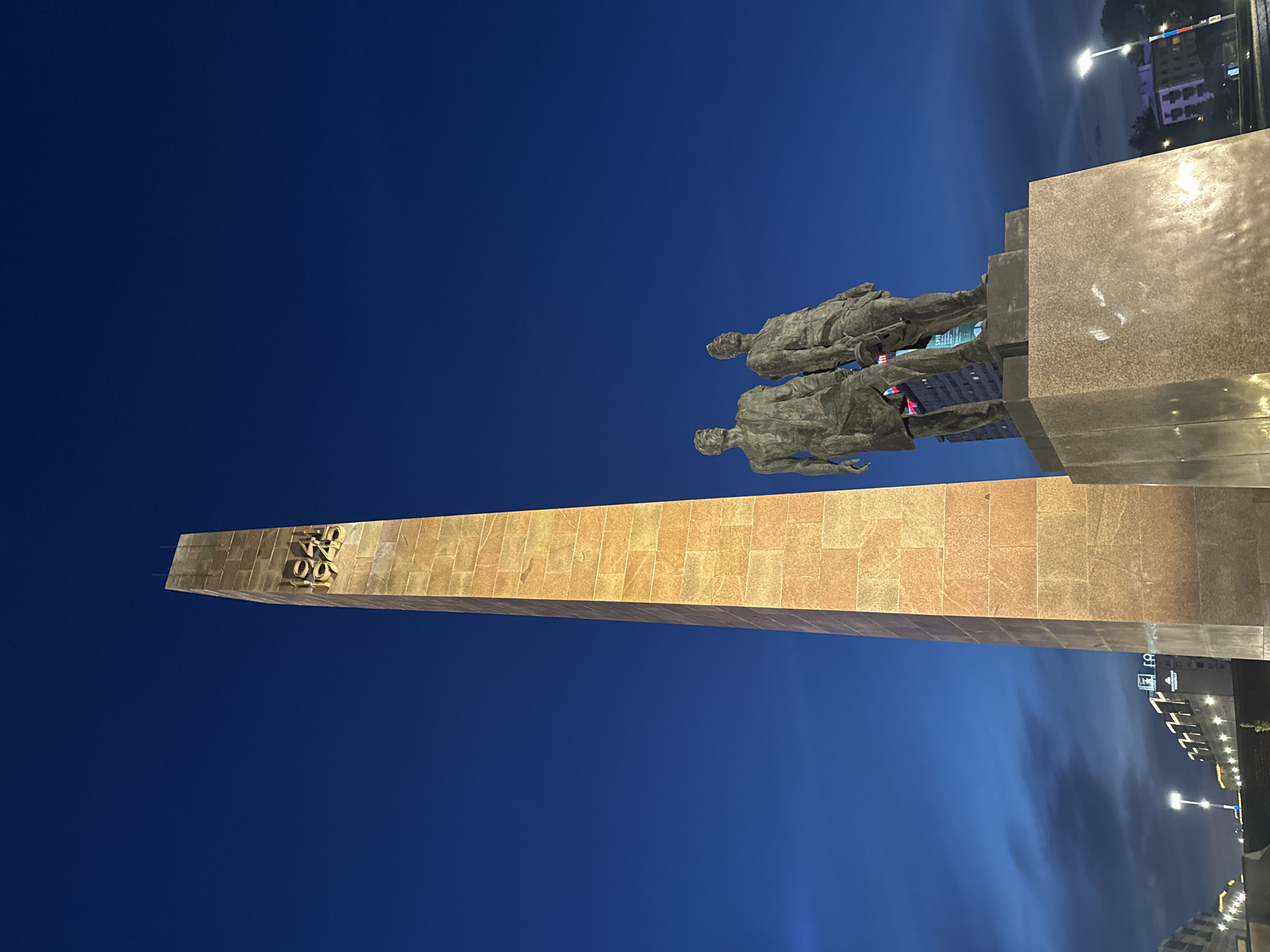 The image displays a monument at night in Saint Petersburg, Russia. The central feature is a tall, rectangular obelisk constructed from light brown granite blocks, tapering slightly towards the top. Near the apex of the obelisk, the golden years "1941" and "1945" are prominently affixed. To the right of the obelisk, on a dark granite pedestal, stand two dark bronze statues of male figures, depicted in heroic, resolute poses, looking towards the right. The figure on the right appears to be holding an object resembling a weapon or tool.
The sky is a deep, rich blue characteristic of twilight or early night, with faint, lighter clouds visible towards the horizon on the left. Artificial lighting illuminates the monument, casting a warm glow on the granite and highlighting the bronze figures. In the background to the left of the obelisk, several buildings are visible, with illuminated windows and signage. One sign distinctly reads "ГОРОД-ГЕРОЙ" (Hero City), and another shows "1945". To the right, streetlights with decorative elements, including what appears to be a Russian flag motif on one pole, line the unseen street, and further architectural structures are visible in the distance. This monument is identified as the Obelisk to the Hero City of Leningrad on Vosstaniya Square in Saint Petersburg.