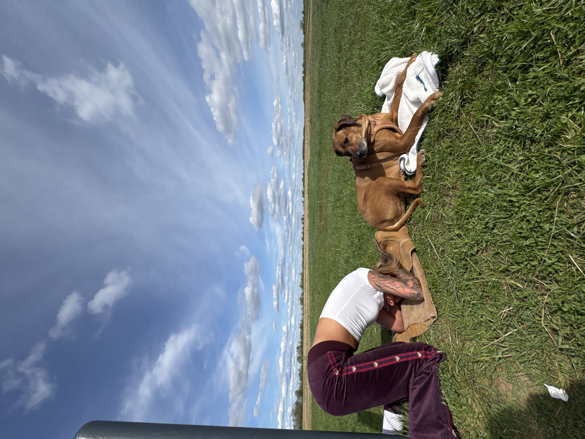 The image shows a woman and a dog outdoors, likely in a grassy field. The woman is kneeling with her head resting on her arms, near a pair of tan-colored boots. She is wearing a white short-sleeved top and maroon sweatpants. Her left arm has tattoos. A brown dog with a harness lies on a white towel next to the woman. The background features a blue sky with clouds and a flat expanse of land. The location is Bol'shoye Kozino, Russia.
