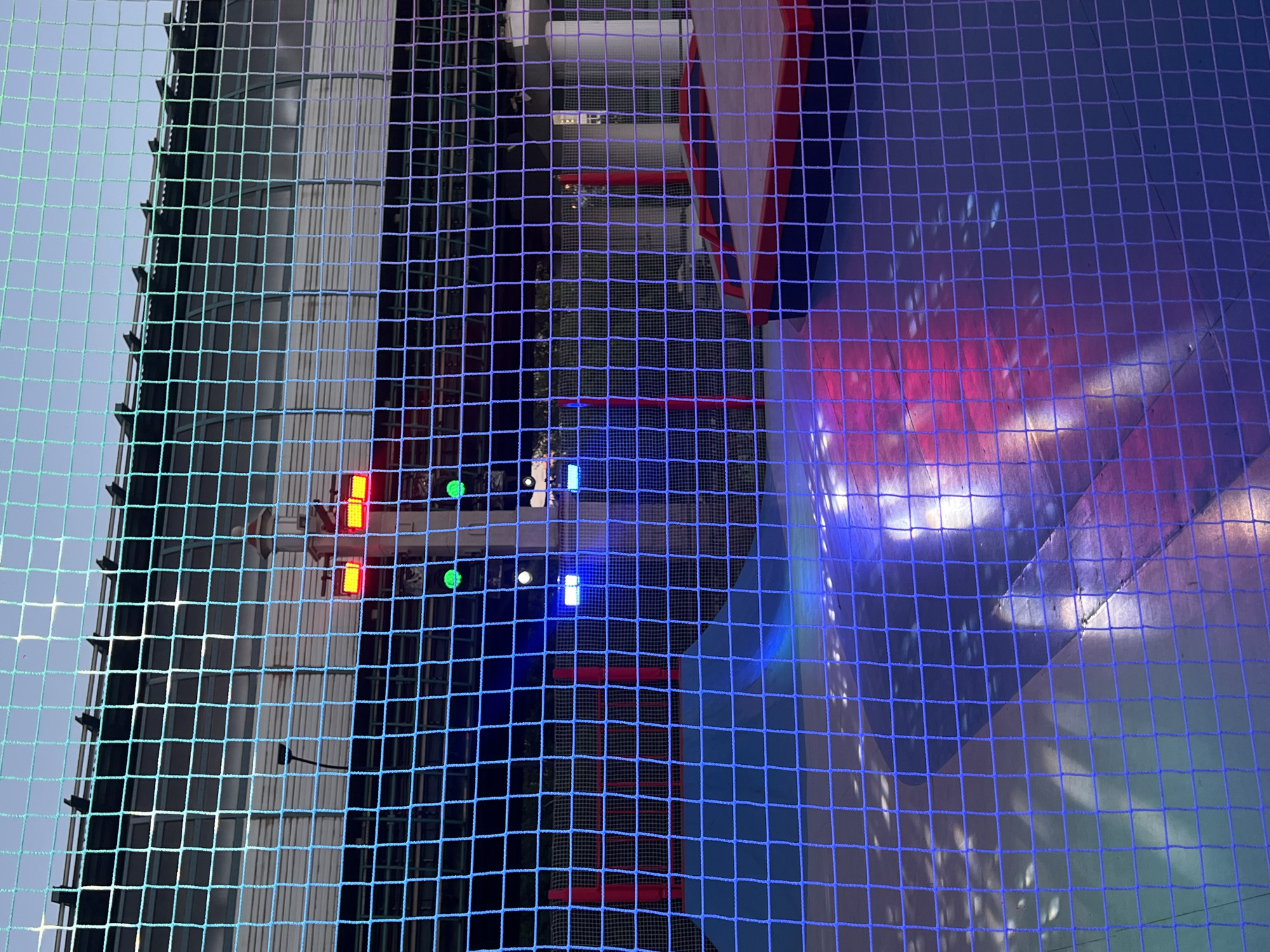 A view from a secured climbing structure in Moscow, Russia. The image is taken from within a climbing net structure, looking towards the adjacent city building. The climbing structure is illuminated with red, green, and blue lights. The background reveals a modern architectural building in Moscow, with windows and structural supports. The scene is indoors.