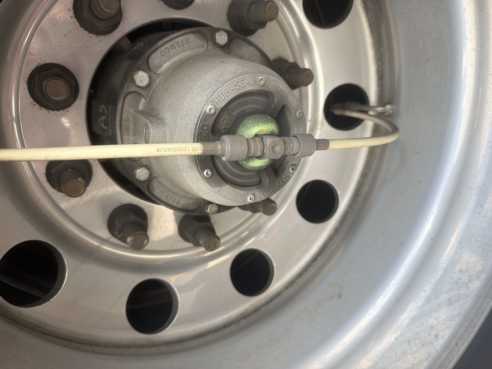 The image shows a close-up view of a truck wheel hub, likely a commercial vehicle. The wheel is made of silver metal and has several circular cutouts. The central hub assembly is also made of metal and has various markings, including "HUB-SEAL" and "STEMCO". There is a green colored cap or seal visible in the center of the hub. A light-colored tube, possibly a tire pressure monitoring system line, extends from the hub towards the viewer. The wheel appears to be attached to a vehicle, and the background suggests an outdoor setting. There are no people or discernible actions taking place in the image. The specific location within the city of Lebanon, United States of America cannot be determined from the image alone.