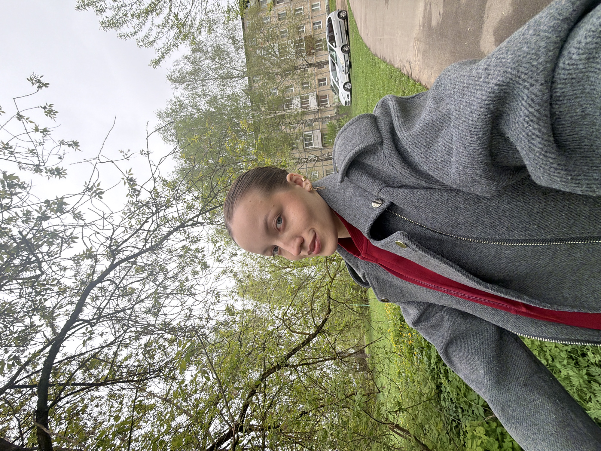 The image shows a woman outdoors in Reshetnikovo, Russia. She is wearing a gray coat over a red top. Behind her are trees with sparse foliage, and in the background is a multi-story apartment building with white cars parked nearby. A paved path is visible along the building's edge.
