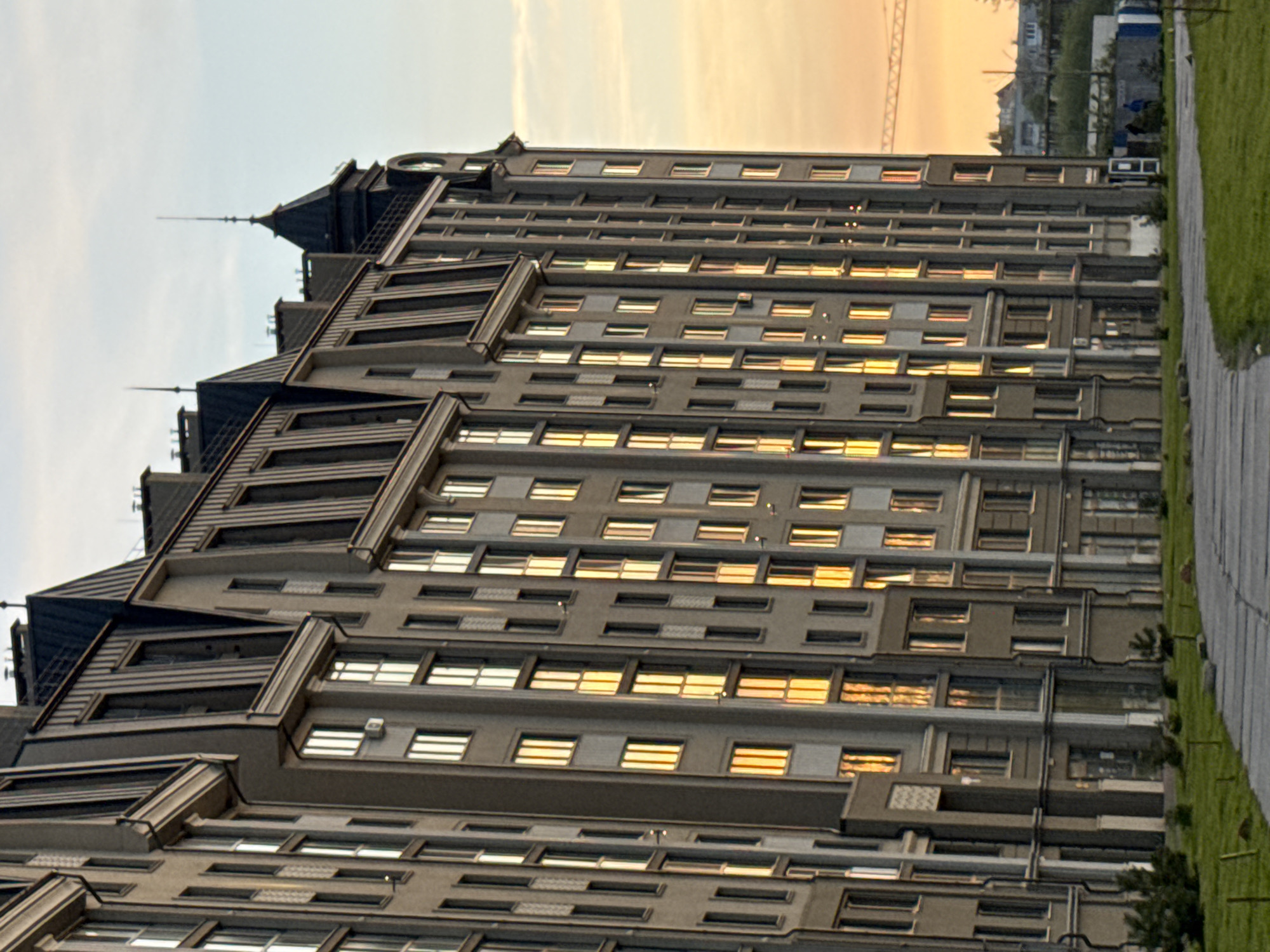 The image shows a multi-story building in Zelenogradsk, Russia. The building has a complex architectural design with multiple gabled sections and spires on top. It features rows of windows reflecting the setting sun. A walkway runs along the right side of the building, bordered by grass. In the background, a glimpse of the sky shows a sunset or sunrise with orange and yellow hues. Further in the background, a crane and other buildings are visible.