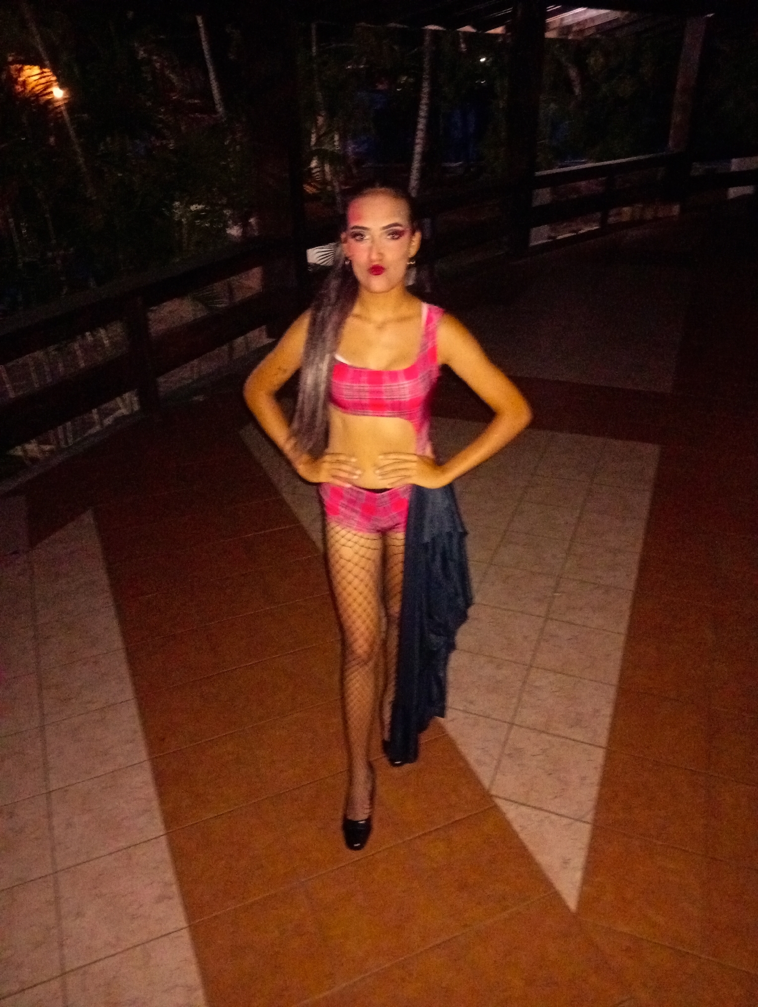 A woman stands on a tiled patio at night in Playa El Agua, Venezuela. She is wearing a pink plaid two-piece outfit, fishnet stockings, and black heels. Her hair is dyed grey, and she wears dramatic makeup. She stands with her hands on her hips, a black garment draped off her right hip. A wooden railing and dense foliage are visible in the background.