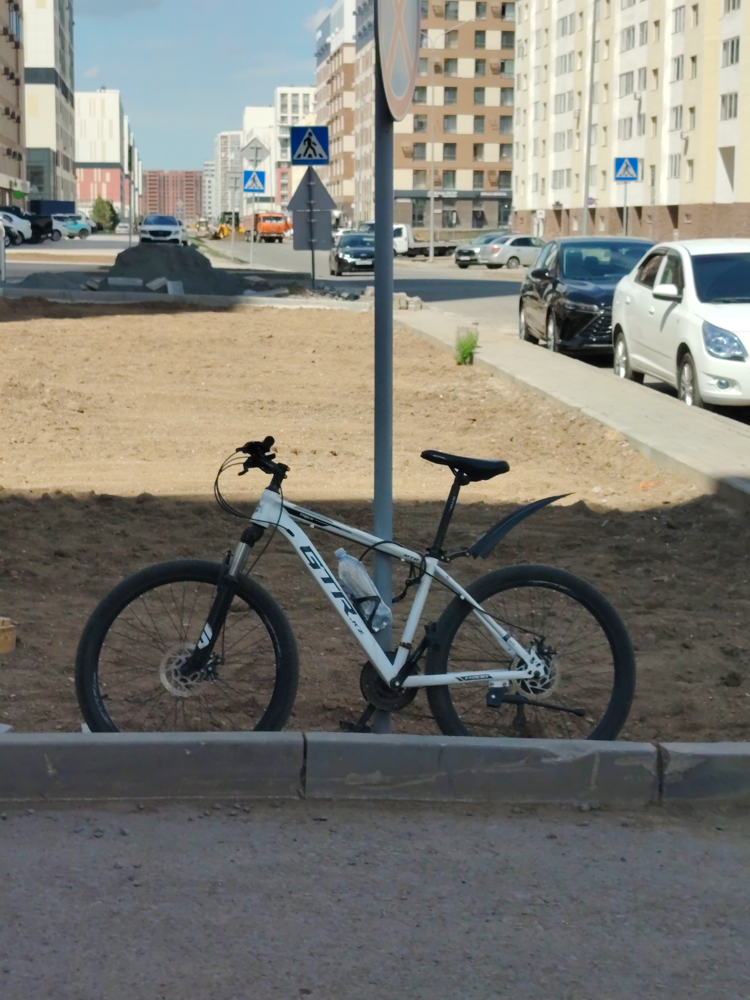 A white mountain bike with the brand name "GTR" on its frame is leaning against a pole. The bike is parked on a dirt area next to a sidewalk. A water bottle is attached to the bike's frame. In the background, several modern apartment buildings are visible, indicating an urban setting. The sky is blue with some scattered clouds, suggesting a clear day. Several cars are parked along the street in the background, and a few traffic signs are also present. One sign displays a pedestrian crossing symbol. Construction materials, such as piles of dirt and sand, are visible on the ground, suggesting ongoing development or roadwork in the area. The scene appears to be taking place during daylight hours, likely late morning or early afternoon, given the bright sunlight. The location is identified as Astana, Kazakhstan. No people are visible in the image.