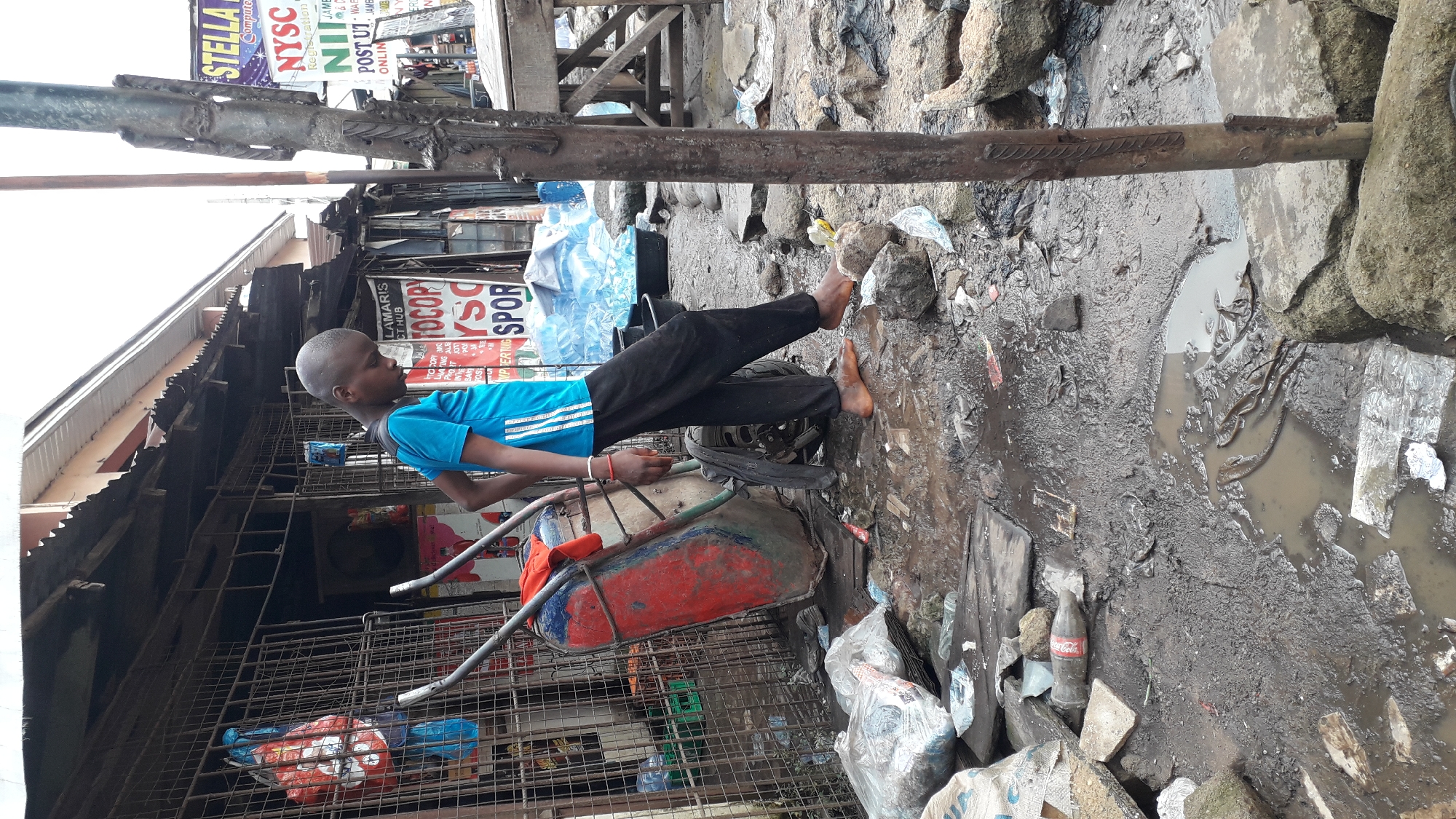 In Onitsha, Nigeria, a young boy stands barefoot in a muddy, debris-strewn street. He is wearing a bright blue t-shirt with white stripes on the sleeves and dark pants. Beside him is a red and blue wheelbarrow, its wheel partially obscured by mud. The background reveals a row of shops with corrugated metal roofs, some featuring signs with writing in English. One sign, partially visible, reads "STELLA" and another reads "NYSC". The ground is covered in puddles, discarded plastic bottles, bags, and various other types of refuse, indicating a generally unkempt urban environment. The overall scene suggests a busy, informal market area with limited infrastructure.