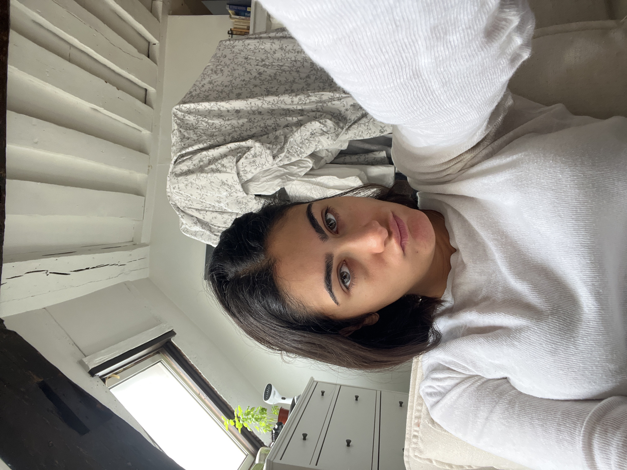 A woman with dark hair and brown eyes is lying on a bed. She is wearing a white sweater. The bed has floral patterned sheets. A white dresser and a window with a plant are visible in the background. The room appears to be an attic space with exposed beams. The location appears to be Paris, France, suggested by the architectural details, which include attic beams.