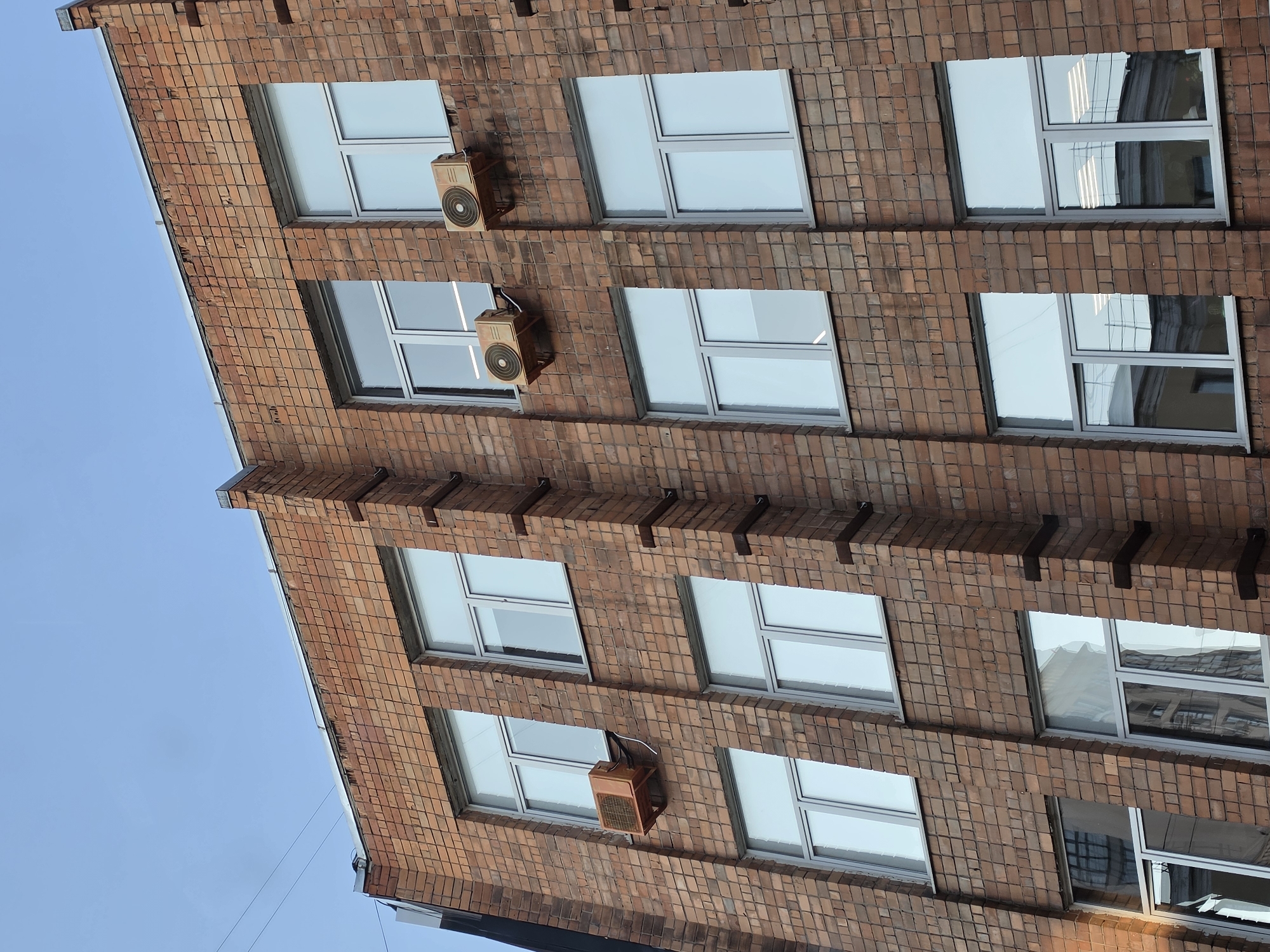 The image shows a close-up, low-angle view of the exterior of a brick building. The building has multiple floors with rows of large, rectangular windows that appear to be made of glass and have silver-colored frames. Some of the windows have interior lighting visible. There are three air conditioning units mounted on the brick facade of the building, two of which are visible in the upper right portion of the image, and one is visible on the left side of the image. The brickwork is reddish-brown and appears to be aged, with some discoloration and texture variations. The sky is clear and blue. The building is likely located in an urban environment given the style of architecture and the presence of air conditioning units. The image does not contain any people or evidence of current activity.