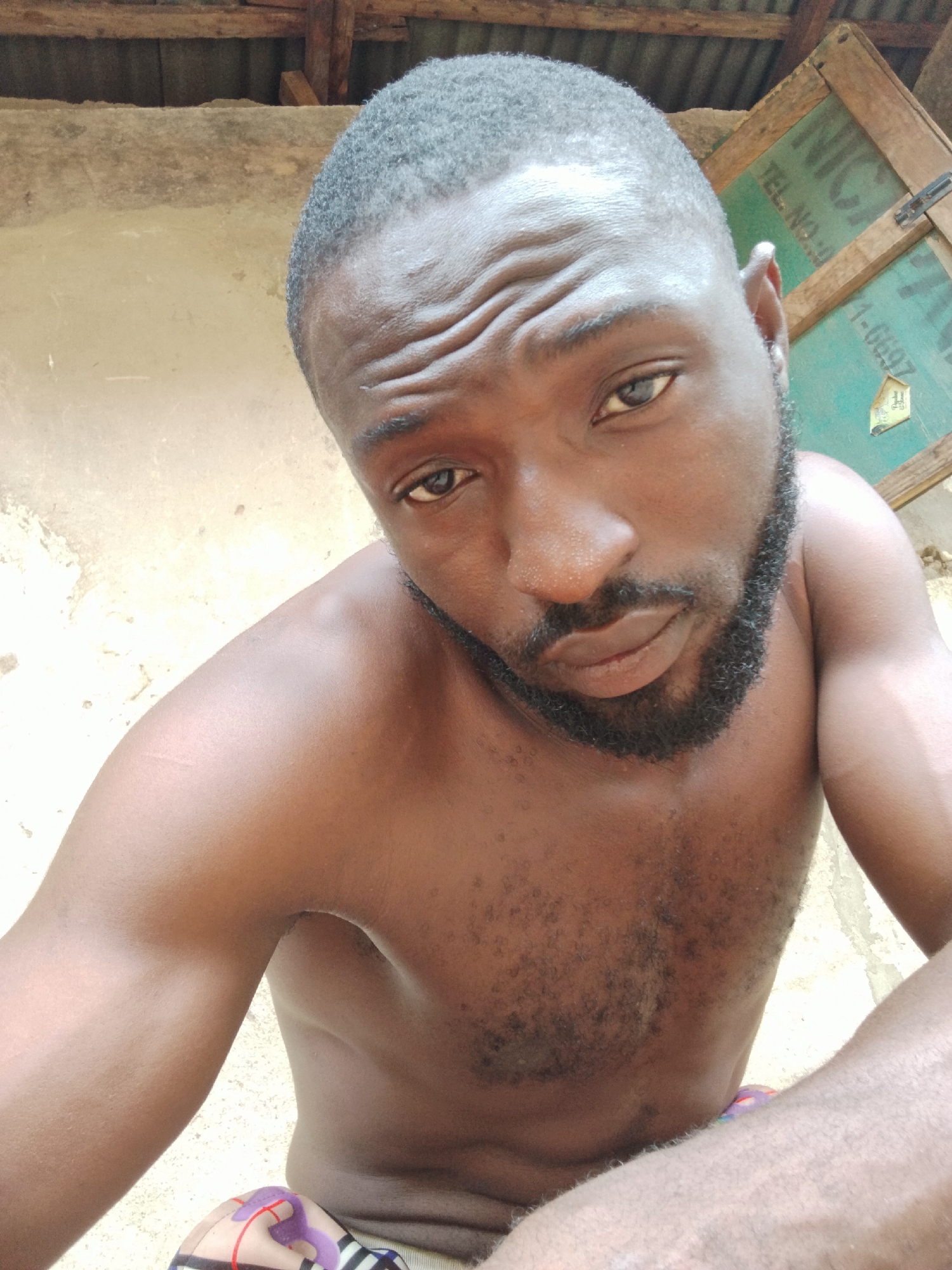 A medium-close up selfie captures a Black man with a dark skin tone, short dark hair, and a full, dark beard and mustache. He is shirtless, displaying a muscular physique and some chest hair. He looks directly at the camera with a neutral expression, showing slight wrinkles on his forehead.
The man's right shoulder and arm are prominent in the foreground, suggesting he is holding the camera. The setting appears to be outdoors in Sabon Tasha, Nigeria, with a rustic background. To the left, there is a light beige or concrete wall. To the right, a wooden structure with a greenish panel is visible, possibly a shutter or door, featuring a metal latch. Above both the wall and the wooden structure, exposed wooden beams and a corrugated metal roof can be seen.
Part of a multi-colored patterned garment, possibly shorts with purple, white, black, and red designs, is visible at the bottom of the frame. A faint, light-colored mark or scar is present on his left shoulder.
Text visible on the green panel, which appears inverted, reads "NIGA" (possibly part of "NIGERIA"), "TEL NO 9", and "1-6997". A small paper tag with unreadable text (possibly "PREMIUM") hangs from the panel. The lighting indicates it is daytime.