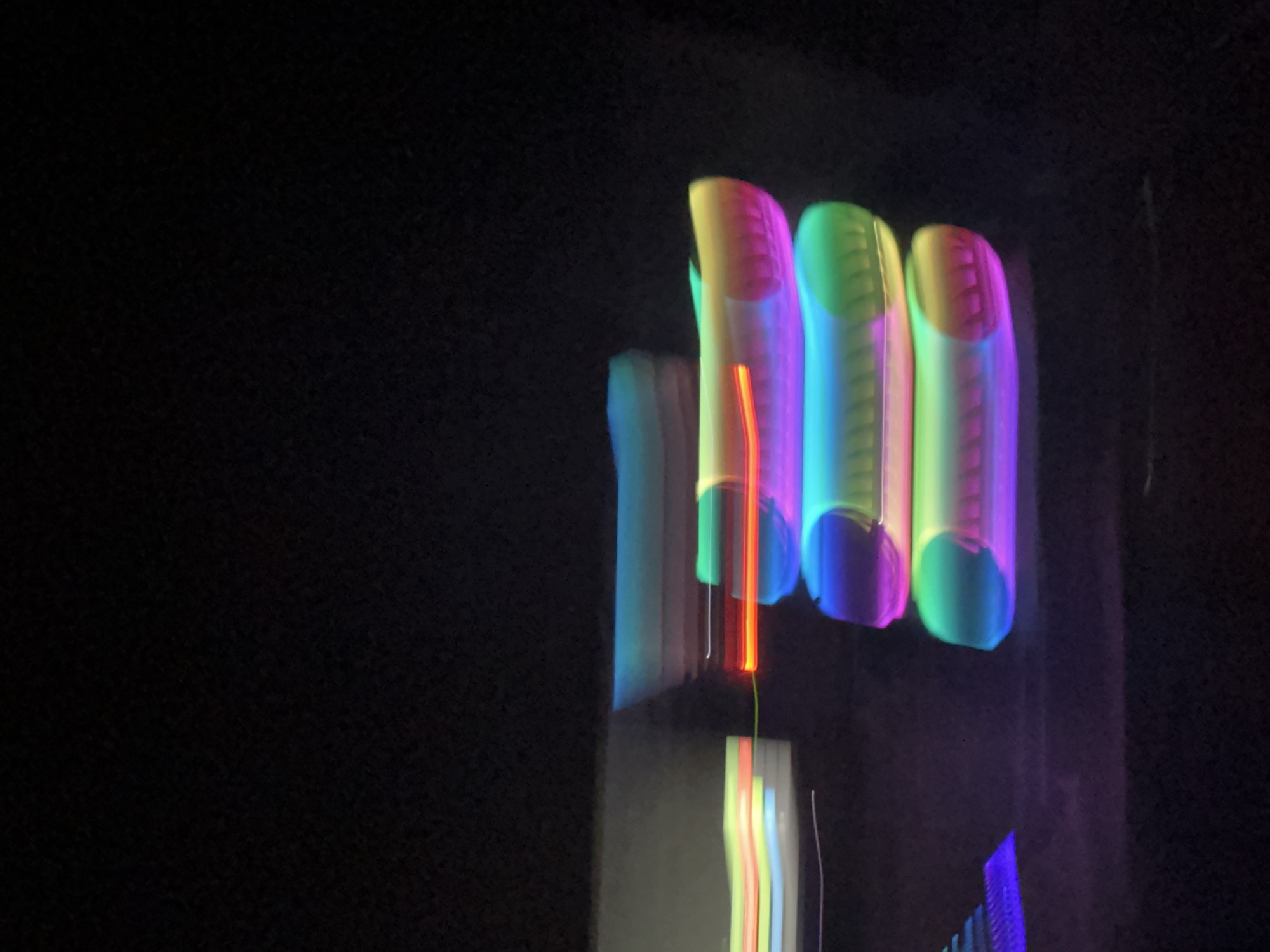 The image is a long exposure photograph of several glowing, cylindrical objects stacked vertically. The objects are illuminated with a spectrum of colors, including blues, greens, yellows, oranges, and purples. The lighting creates streaks and blurs due to motion during the exposure. The background is predominantly black, suggesting a dark or nighttime environment. On the left side of the image, there are faint, horizontal streaks of light in various colors, possibly indicating other moving lights or objects out of focus. The overall impression is abstract and artistic, with a focus on light and color trails. This photograph was taken in Stallings, North Carolina, United States of America.