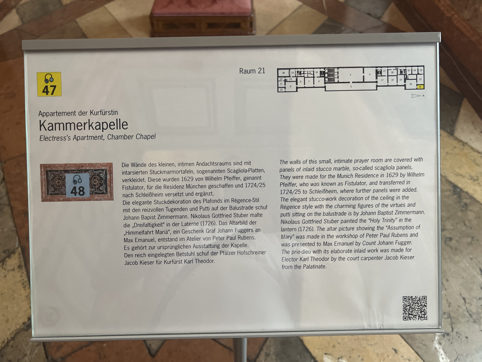 The image shows an informational plaque displayed in Oberschleißheim, Germany, within the New Schleissheim Palace. The plaque is positioned in front of an exhibit or room, featuring a floor plan labeled "Raum 21" (Room 21) at the top. The plaque contains text in both German and English, describing the "Appartement der Kurfürstin" (Electress's Apartment) and its "Kammerkapelle" (Chamber Chapel). Below the title is a small square image with a number "48" inside a headphone symbol, and an ornate patterned border. The text details the construction and decoration of the chapel, mentioning artists like Wilhelm Pfeiffer, Johann Baptist Zimmermann, and Peter Paul Rubens. The environment appears to be an indoor exhibition space with a polished floor. The plaque itself is framed and appears to be made of a reflective material.